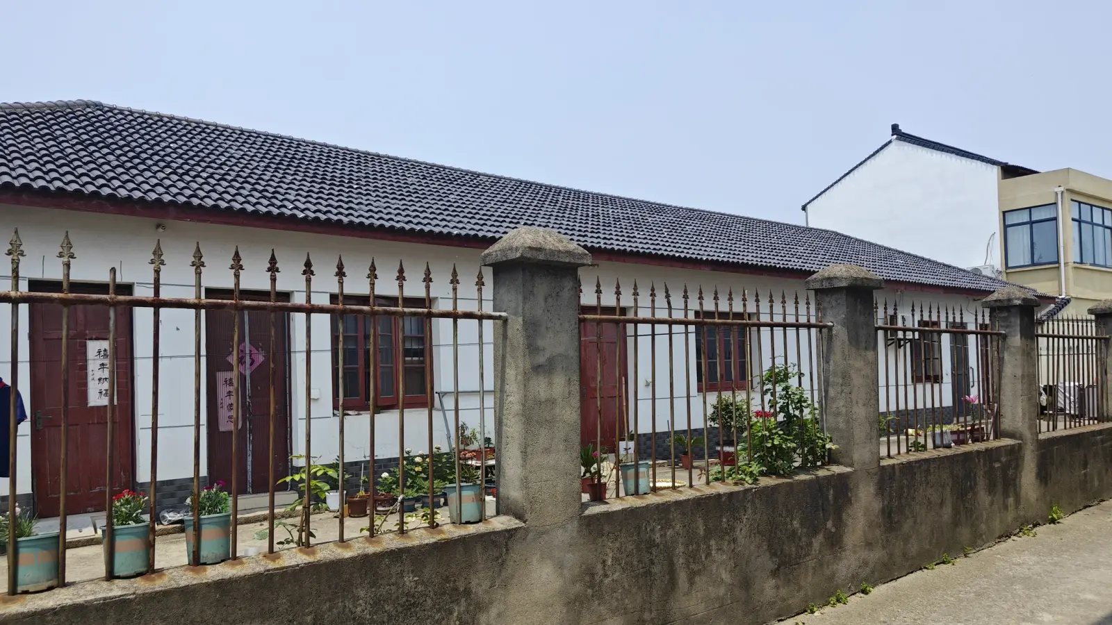 A whitewashed building with a long grey tile roof seen along a stone-posted iron fence with potted plants.