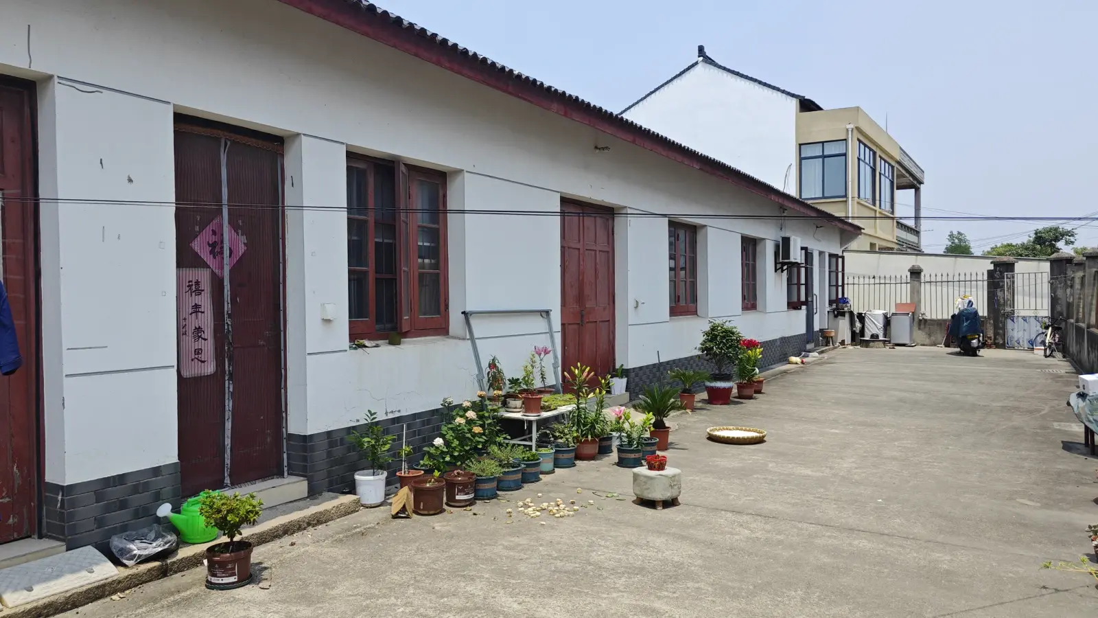 A whitewashed building with dark red doors and windows, with potted flowers along the paved yard.