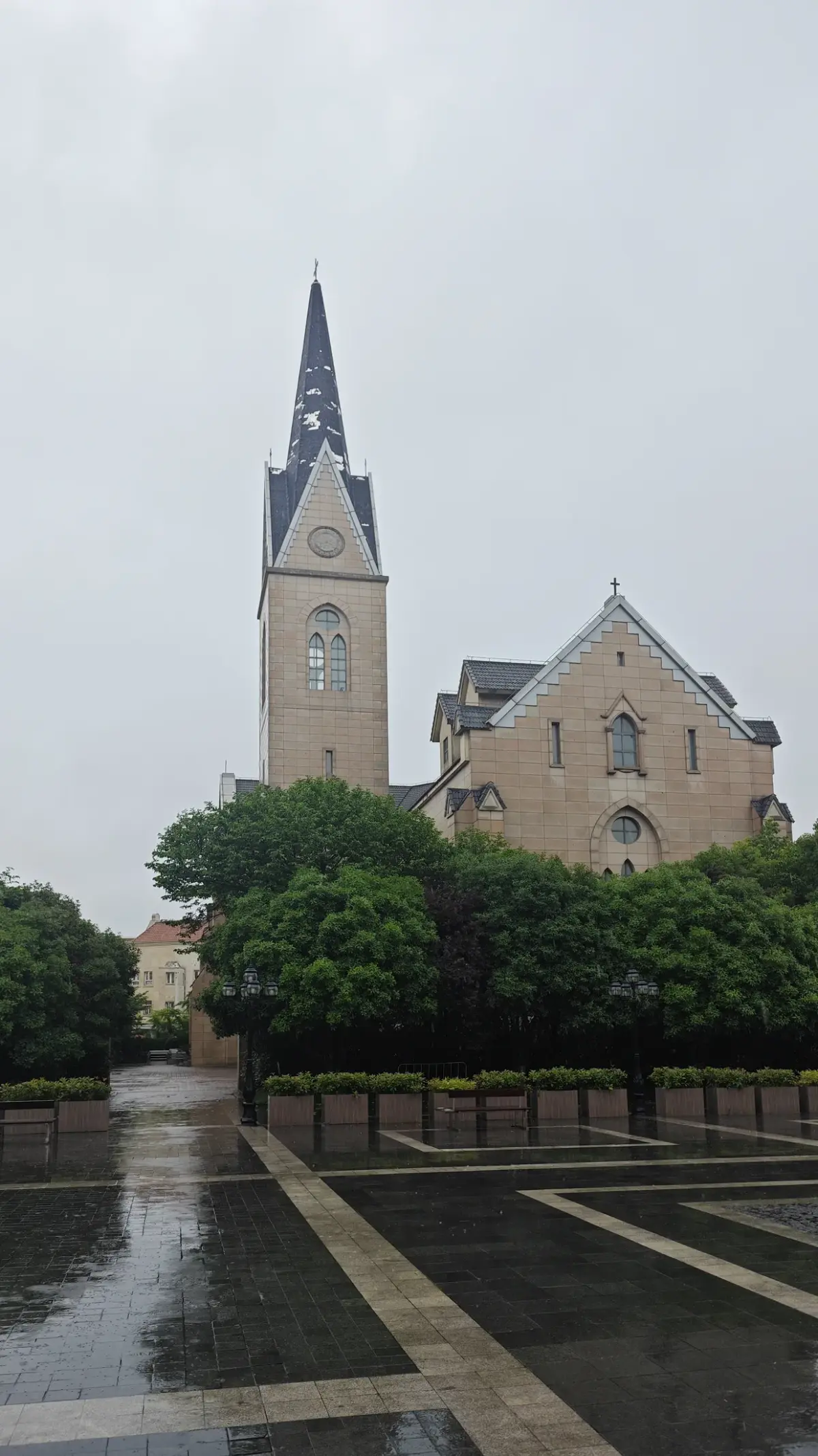 A pale stone Gothic church with a tall clock tower topped by a dark slate spire and a gabled front, above green trees and a wet paved plaza.