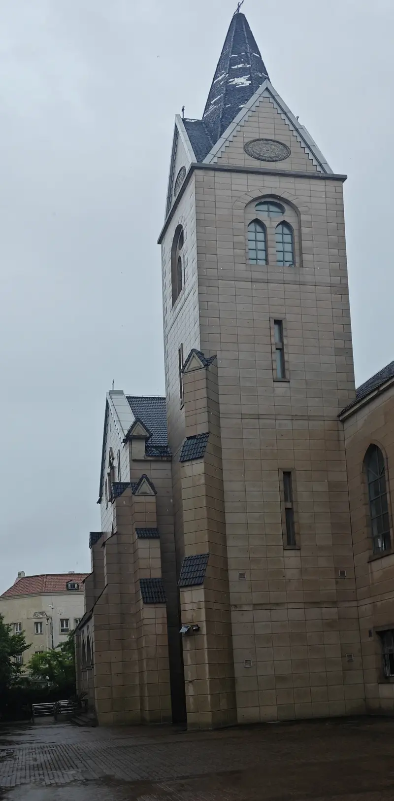 Looking up at a stone church tower with arched belfry windows and a slate spire, beside a long side wall with tall pointed windows.