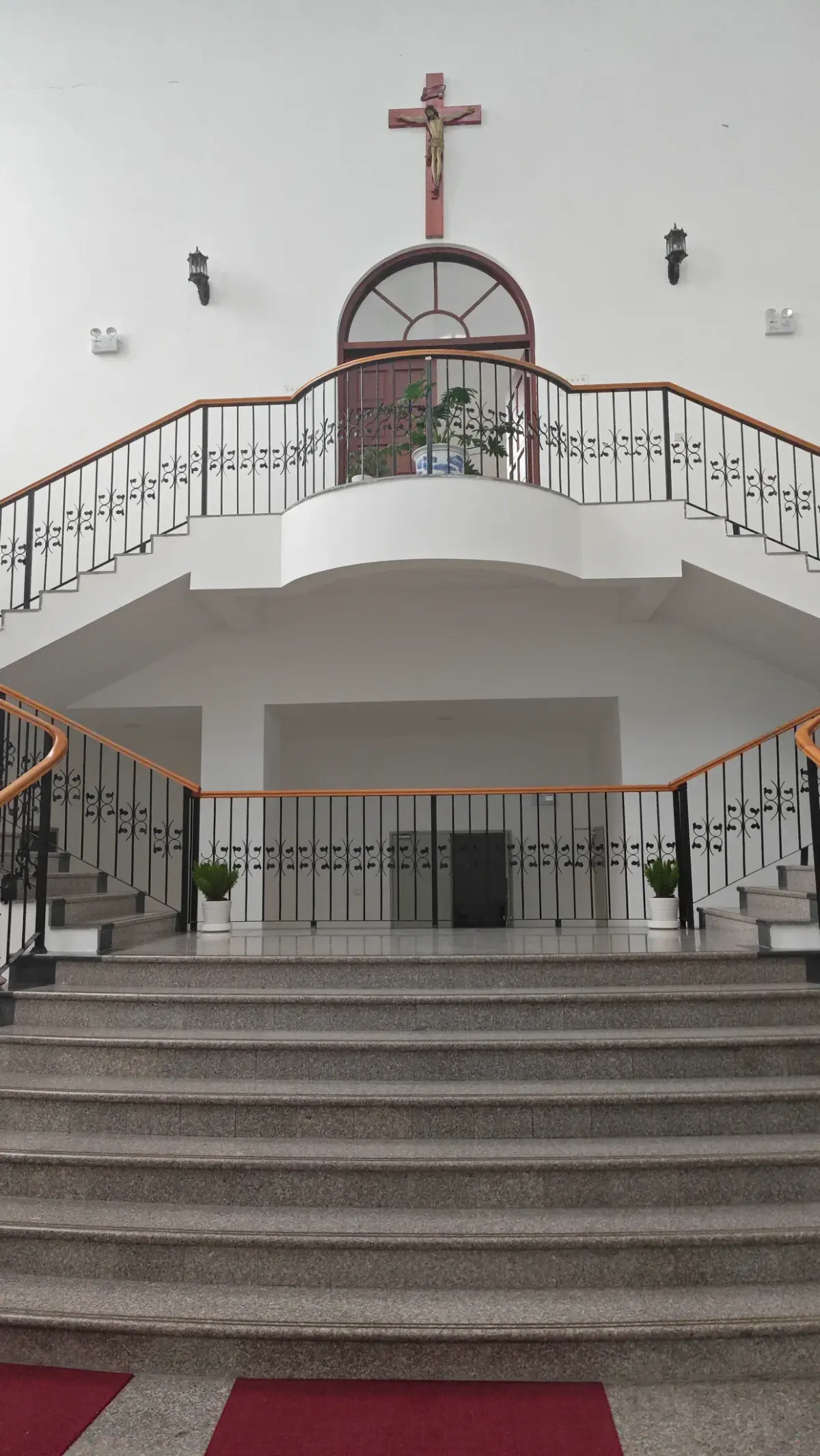 A broad flight of granite steps leading to a curved balcony with railings, and a large crucifix above an arched window on a white wall.