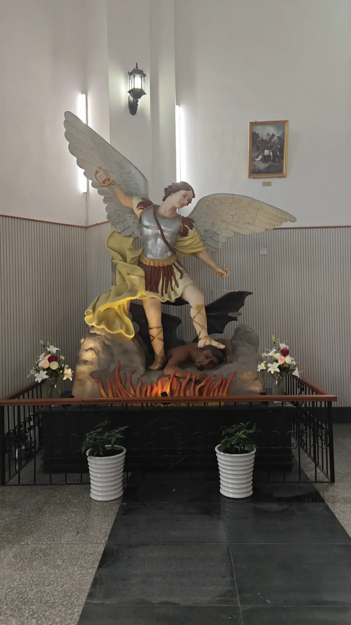 A colourful winged statue of a warrior angel standing over a dark figure and red flames, flanked by white flower pots.
