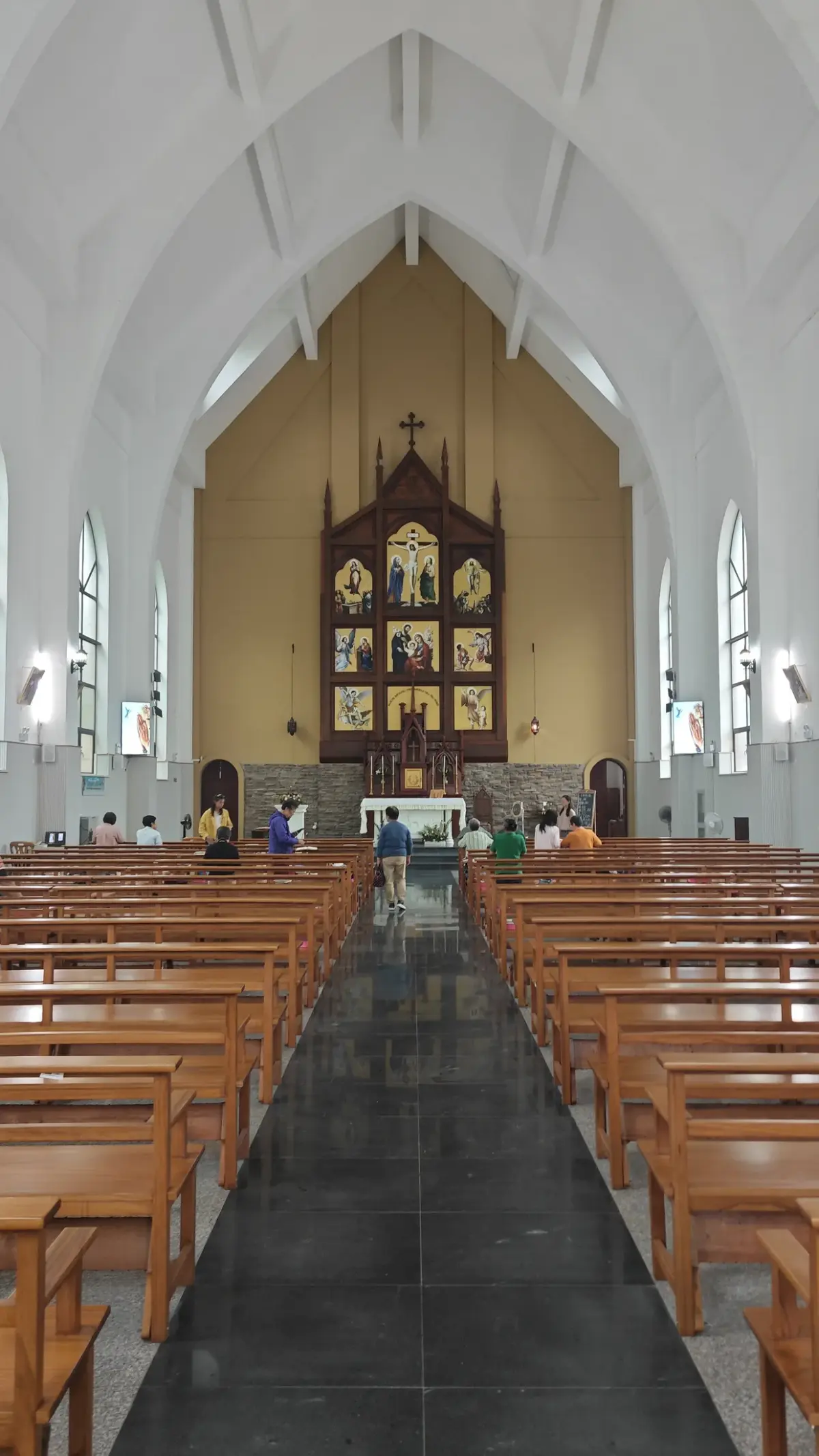 A tall church nave with white roof trusses, rows of pale wooden pews and a dark carved altarpiece on a golden-yellow end wall.