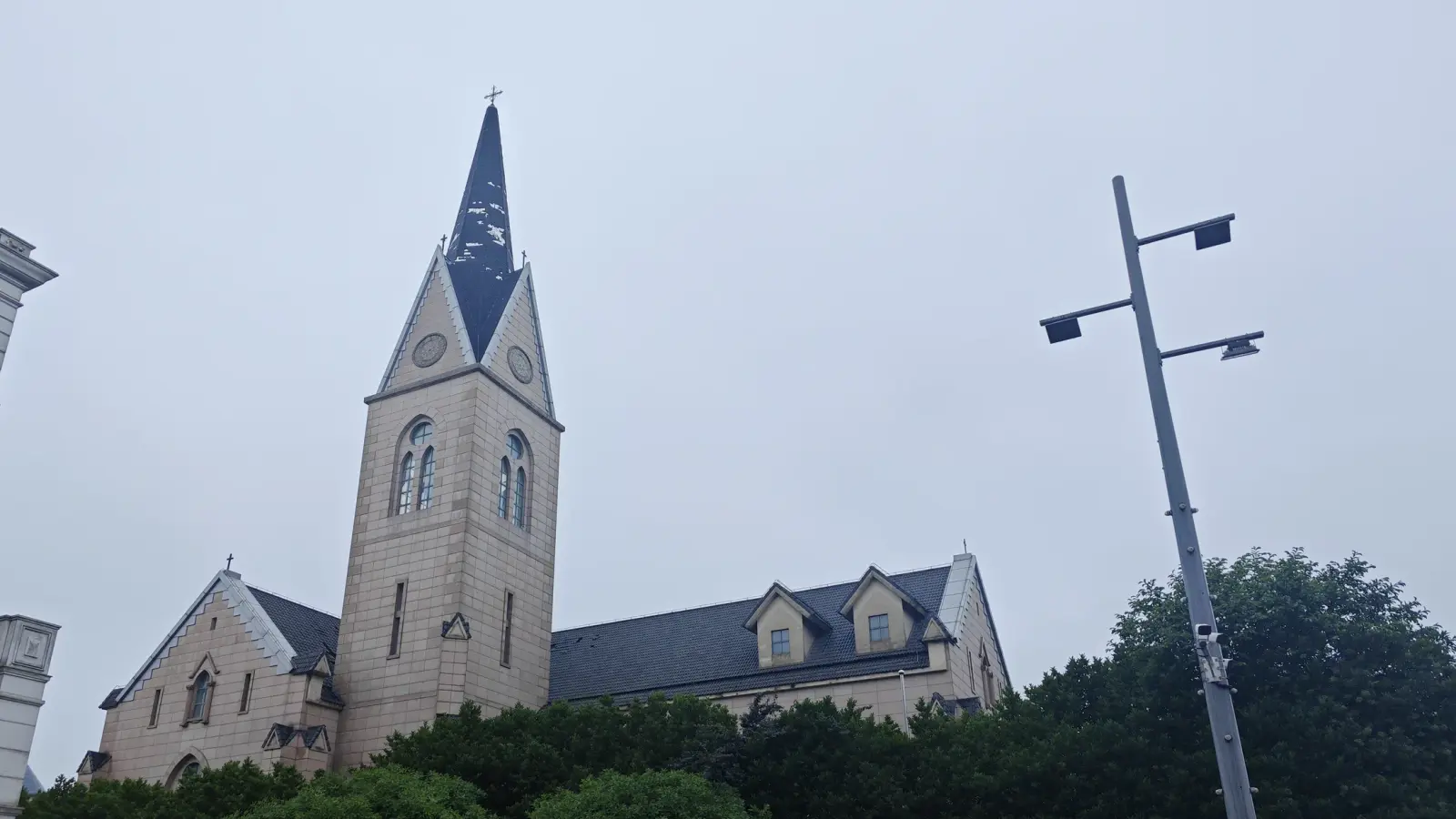 A stone church tower with a dark slate spire and a roof with gabled dormers, against a pale sky, with a lamp post at right.
