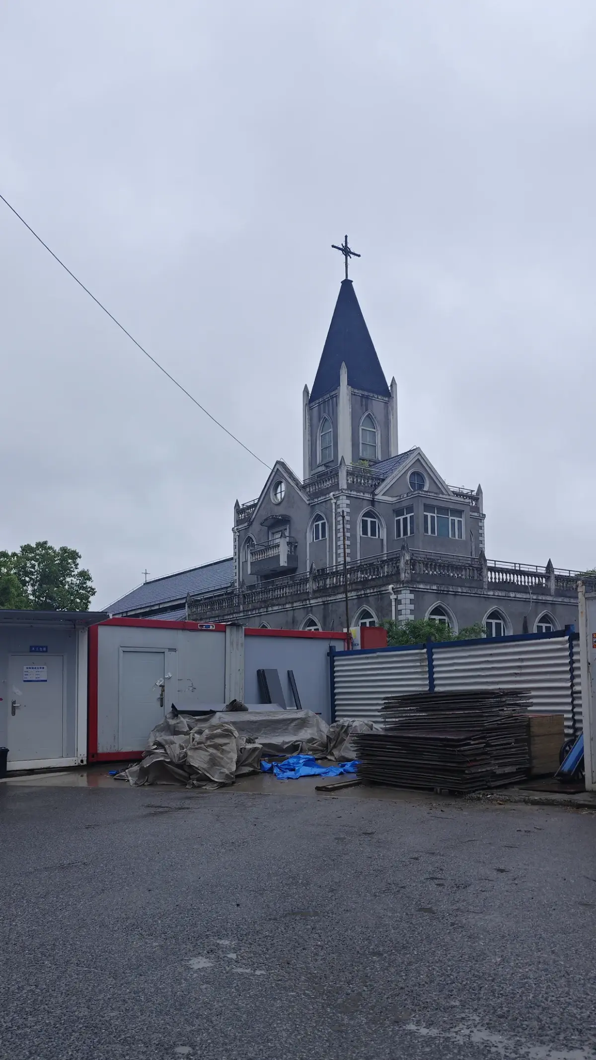 A grey stone church with a gabled front and a tall dark spire, seen across a wet car park with portable site cabins.