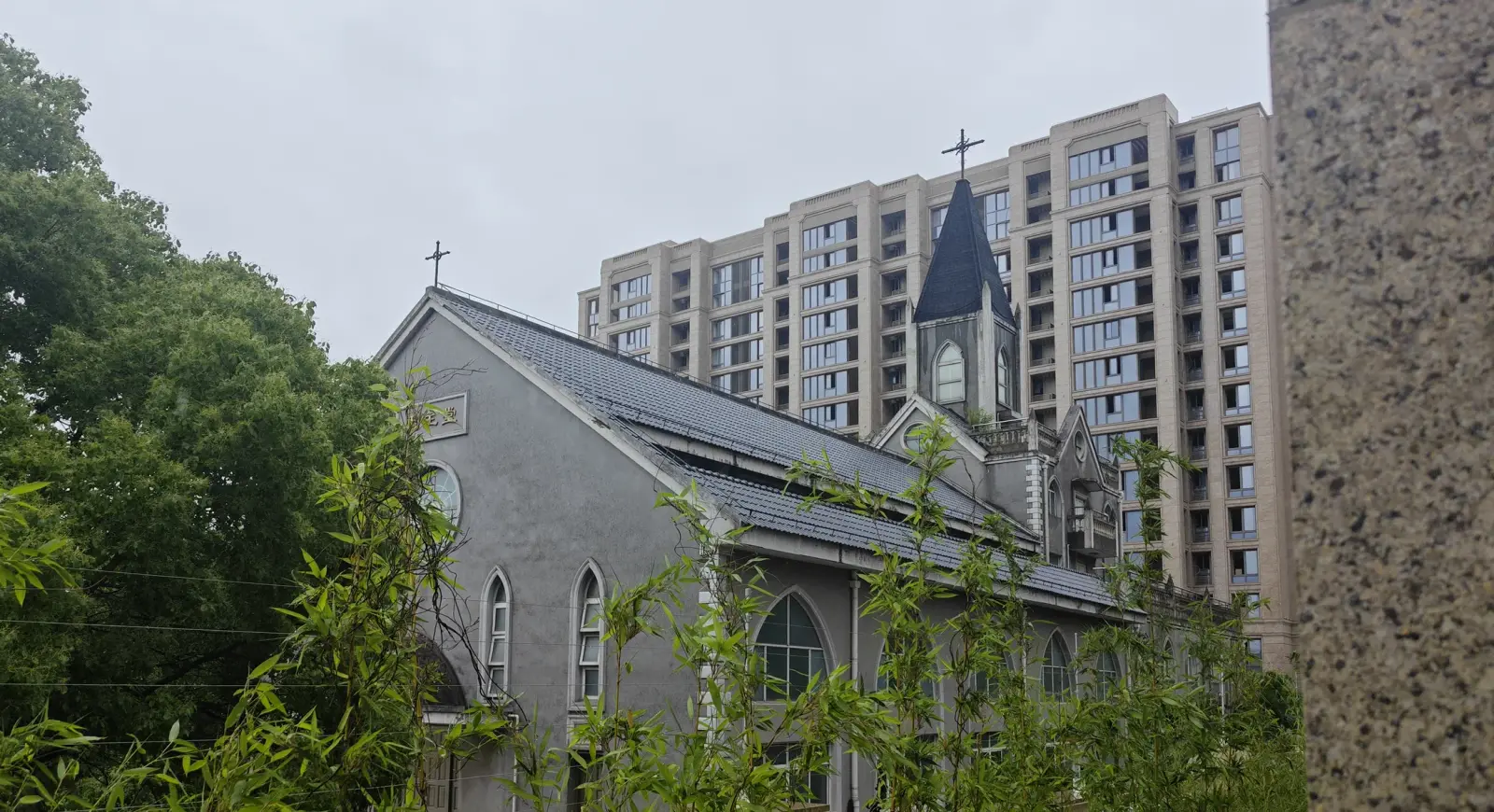 A grey church with a pitched roof, lancet windows and a slim dark spire with a cross, with a tall apartment block behind it.