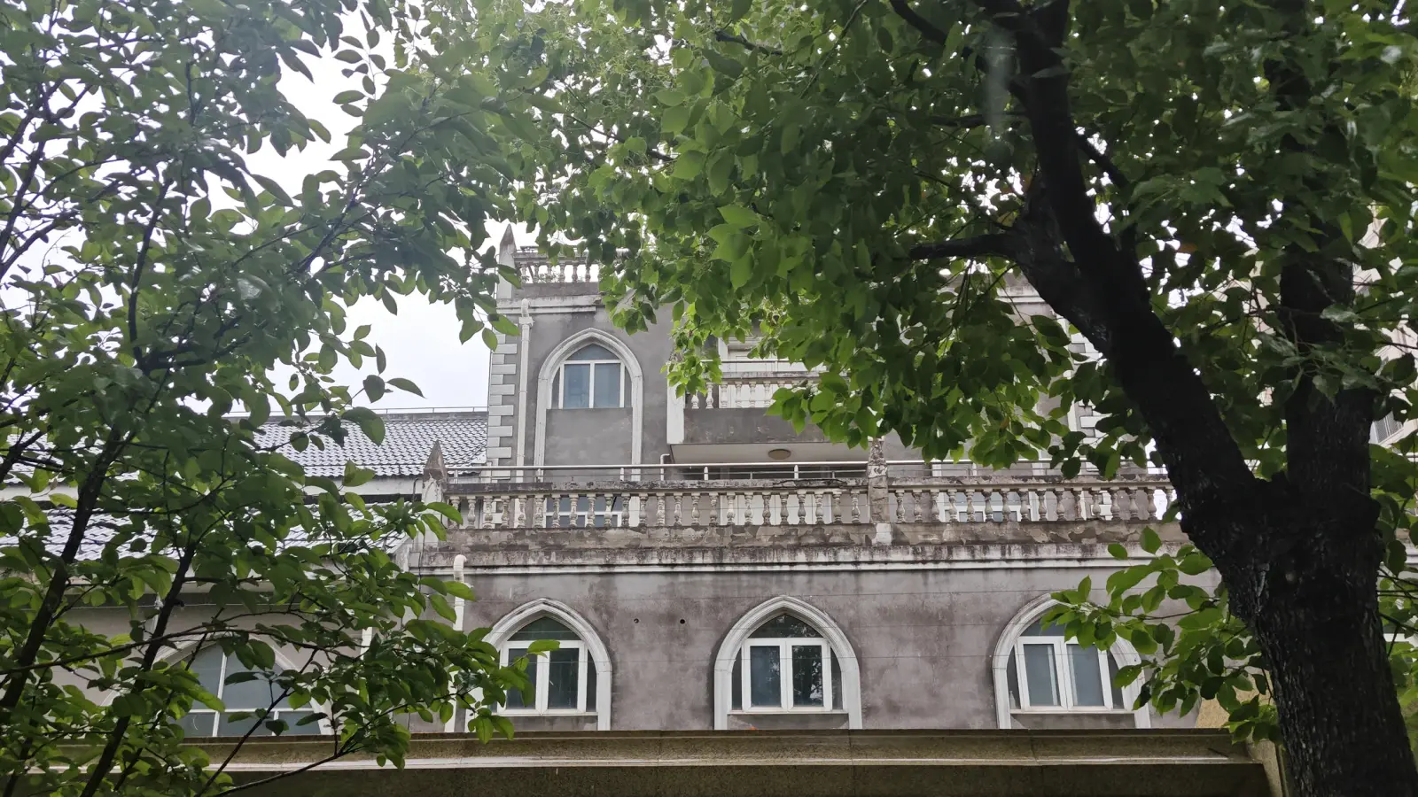 Pale grey walls with white-framed pointed windows and a stone balustrade, seen through tree branches.
