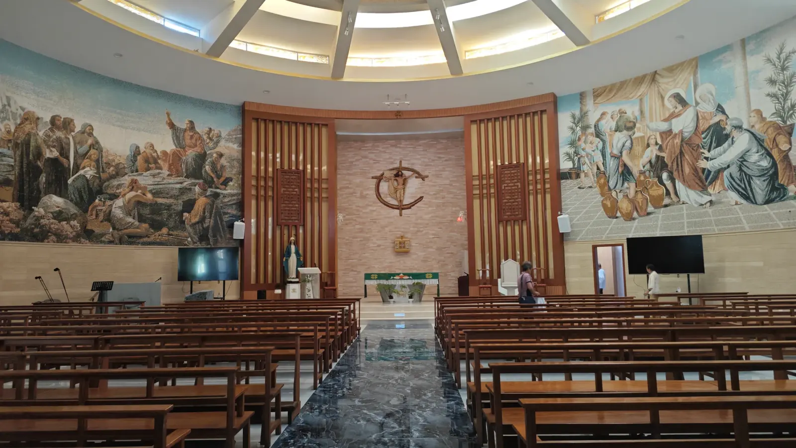 A church interior with long wooden benches, a green marble aisle, a cross behind the altar and painted mosaics on the side walls.