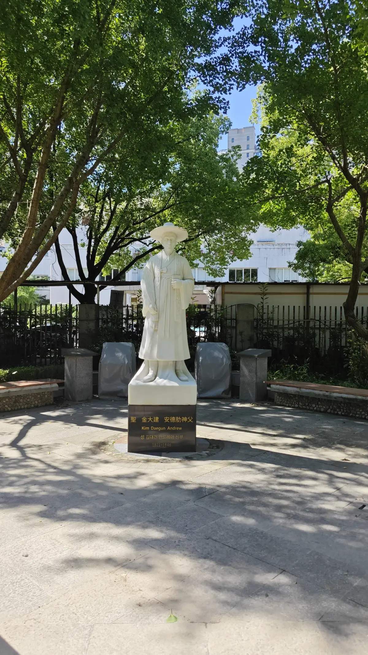A white statue of a robed figure wearing a broad hat on a stone base, under trees.