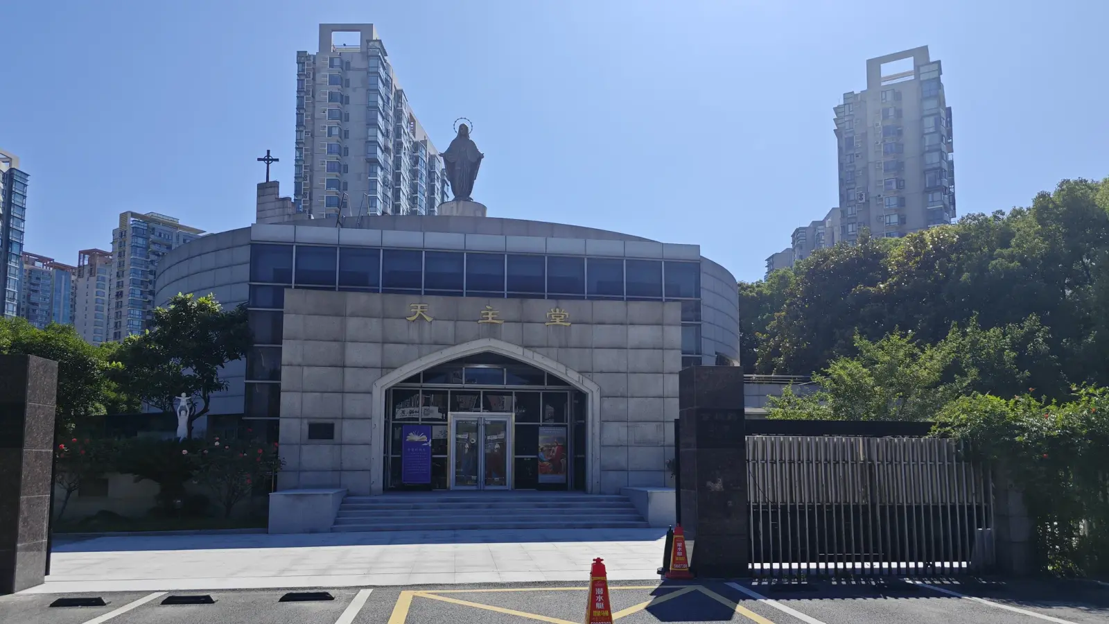 A modern church with a rounded stone wall, a pointed-arch entrance, a cross and a statue on the roof, with apartment towers behind.