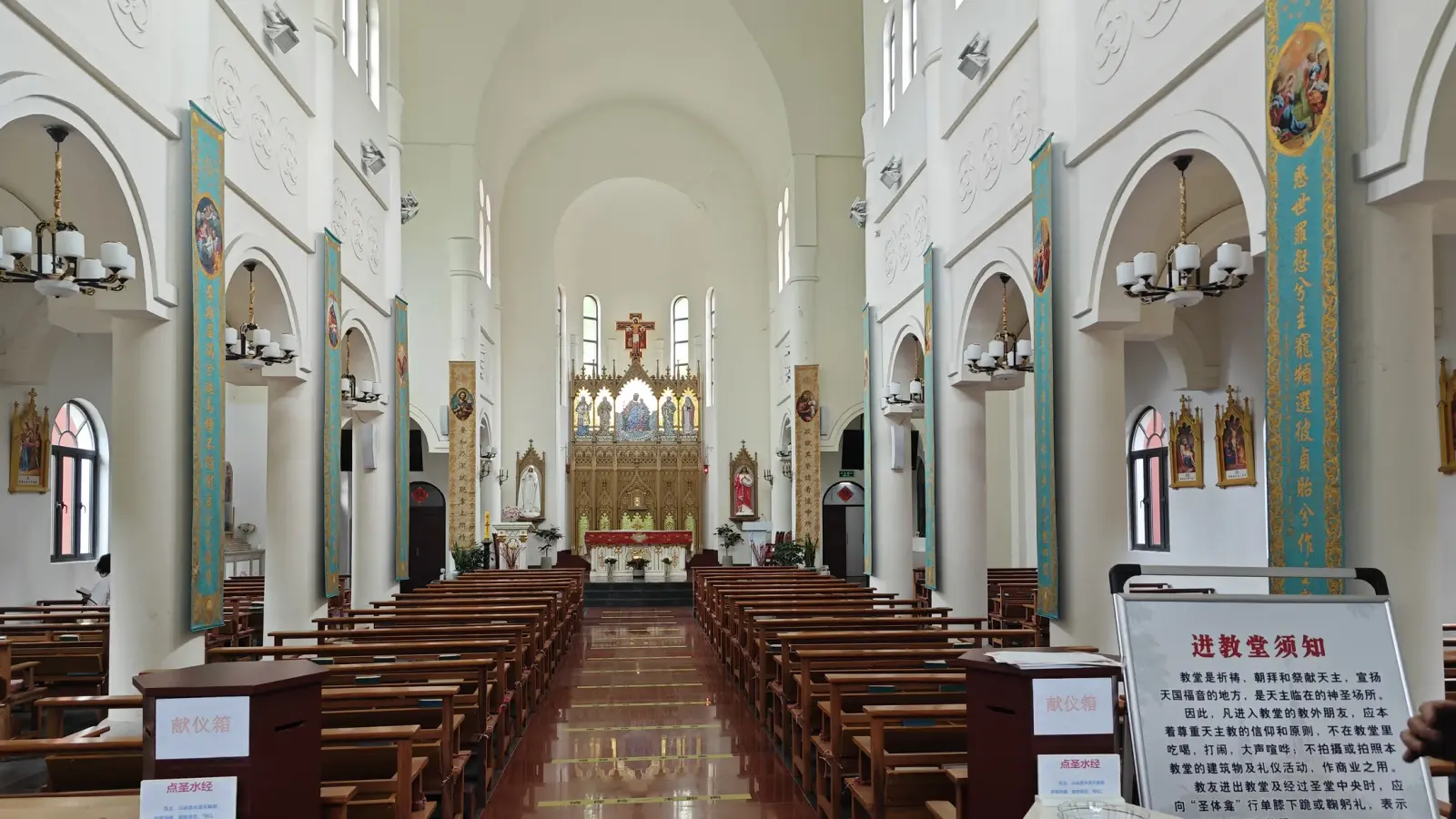 A tall white church interior with round arches, hanging chandeliers, banners, red-brown tiled aisle and a gold altarpiece at the end.