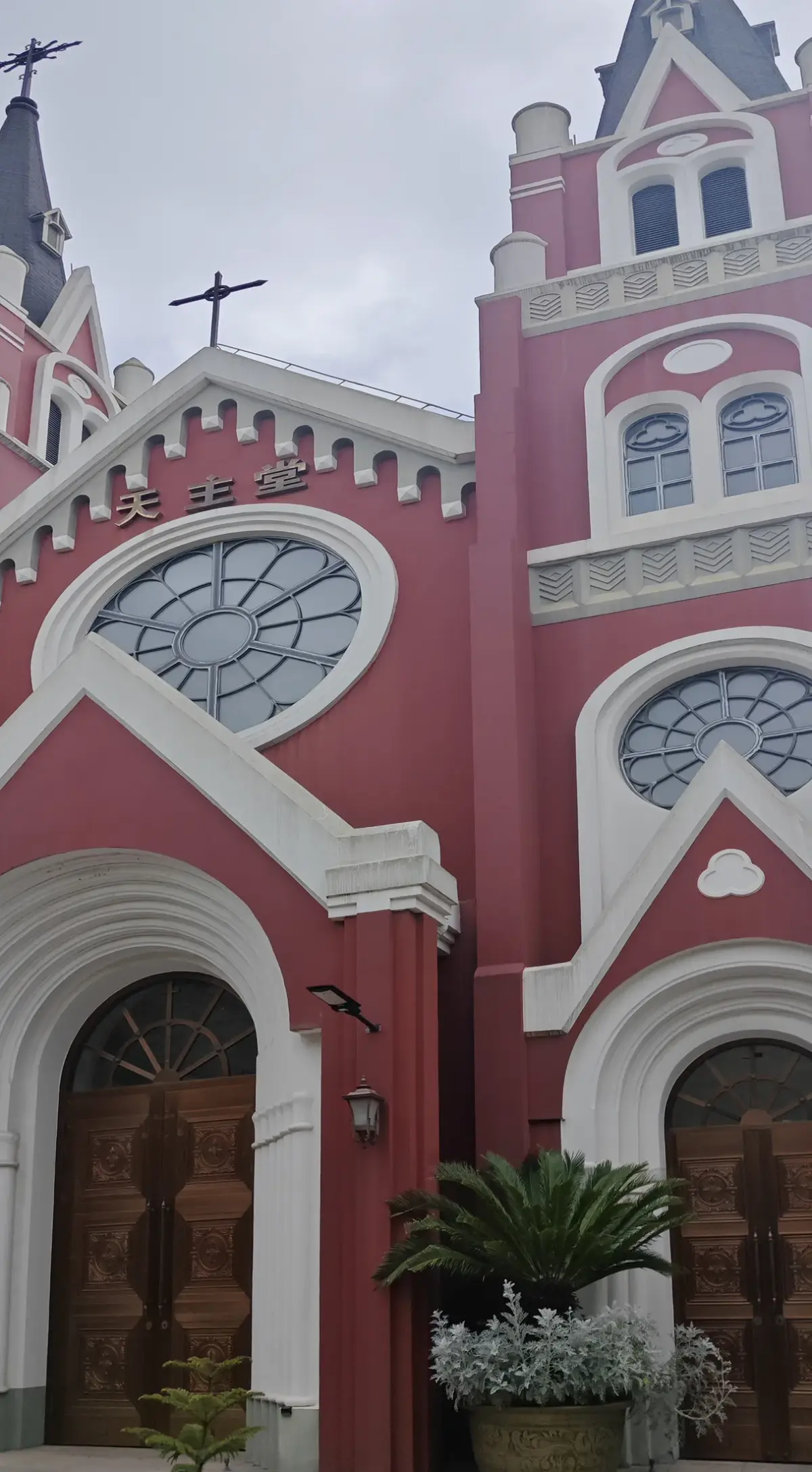 A close view of a red church front with white-framed round windows, a pointed gable, tall arched wooden doors and a spire.