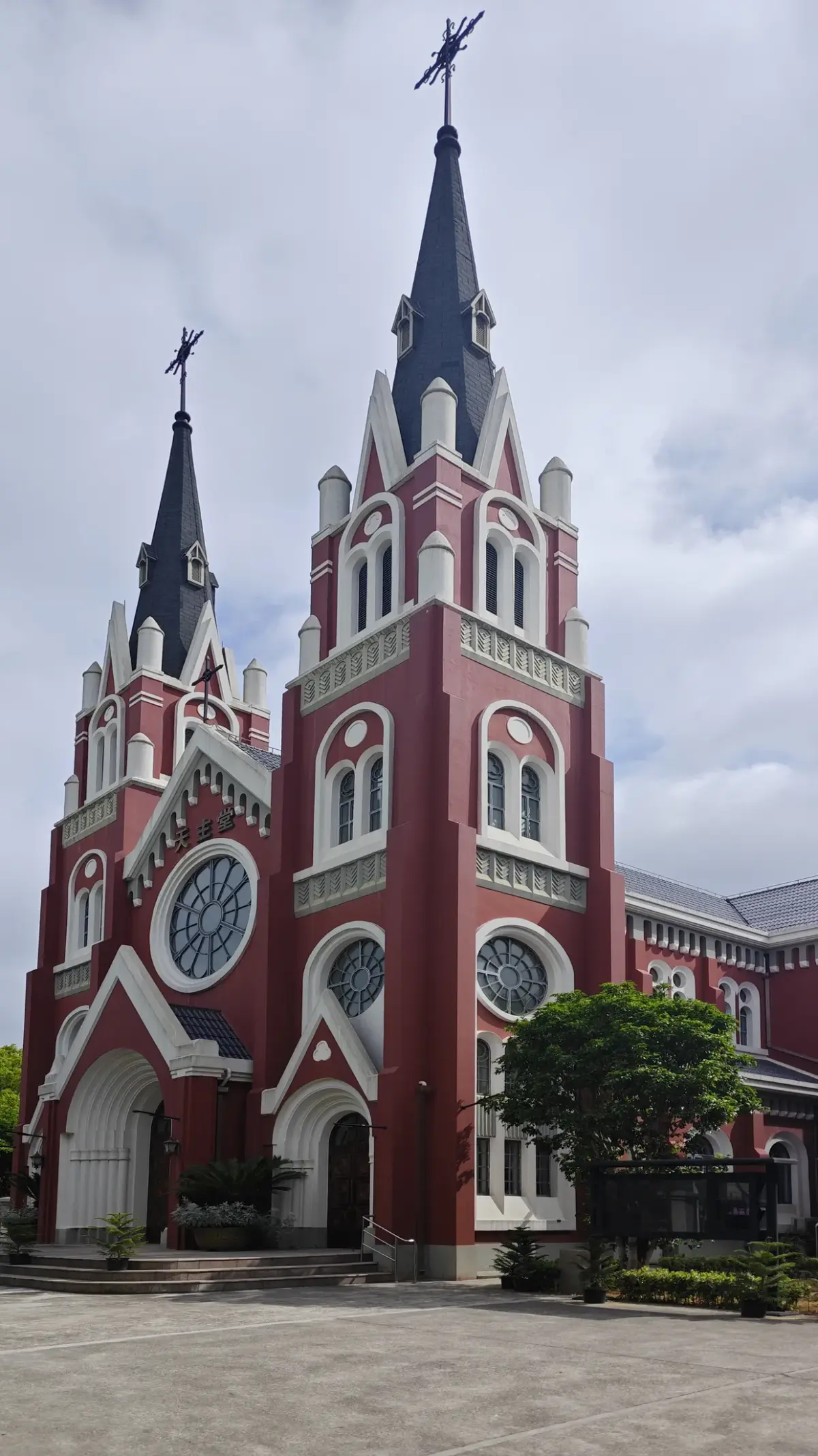 A red church front with two towers topped by dark slate spires and crosses, round windows and white arched doorways, under a cloudy sky.