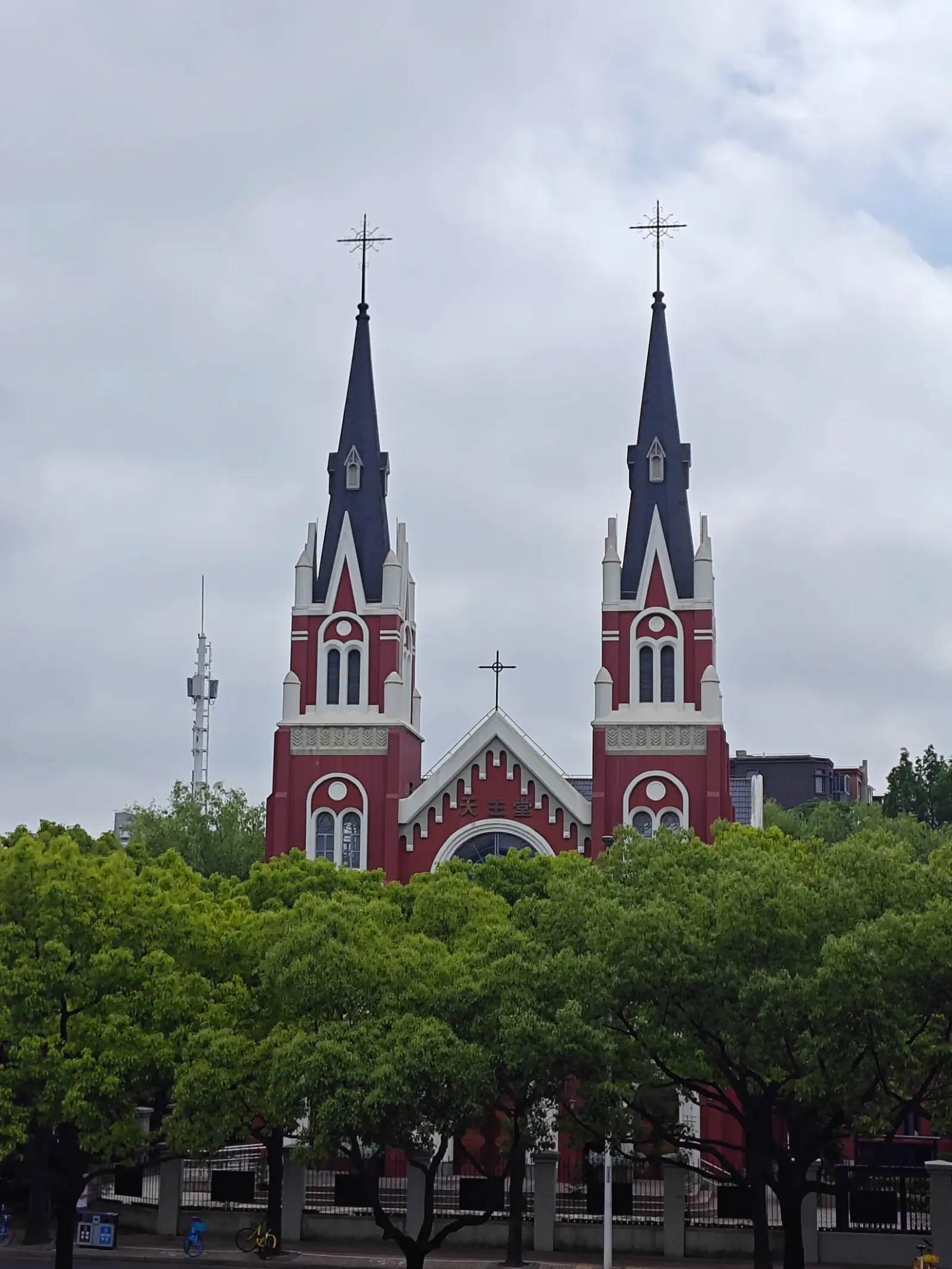 Two red towers with dark pointed spires and crosses and a pointed central gable, seen over a row of trees under a pale sky.