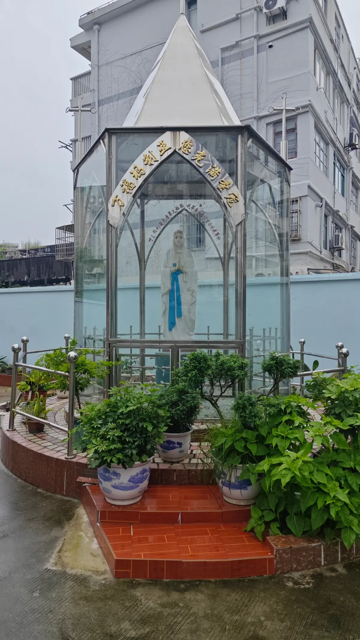 A statue of a woman in blue inside a hexagonal glass shrine with a white pointed roof, among potted plants.