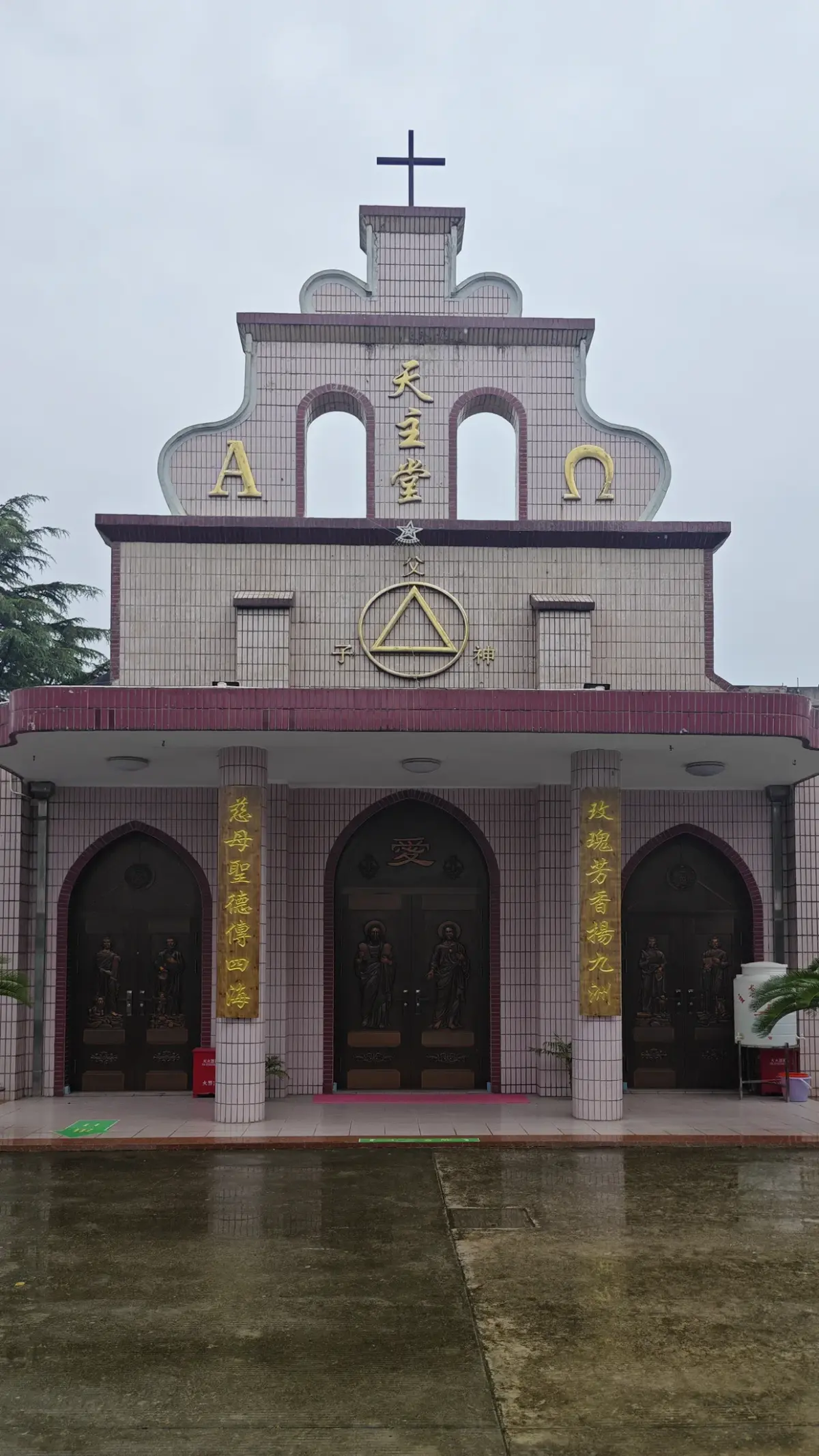 A pink tiled church front with a stepped gable topped by a cross, Greek letters Alpha and Omega, and three arched wooden doors.