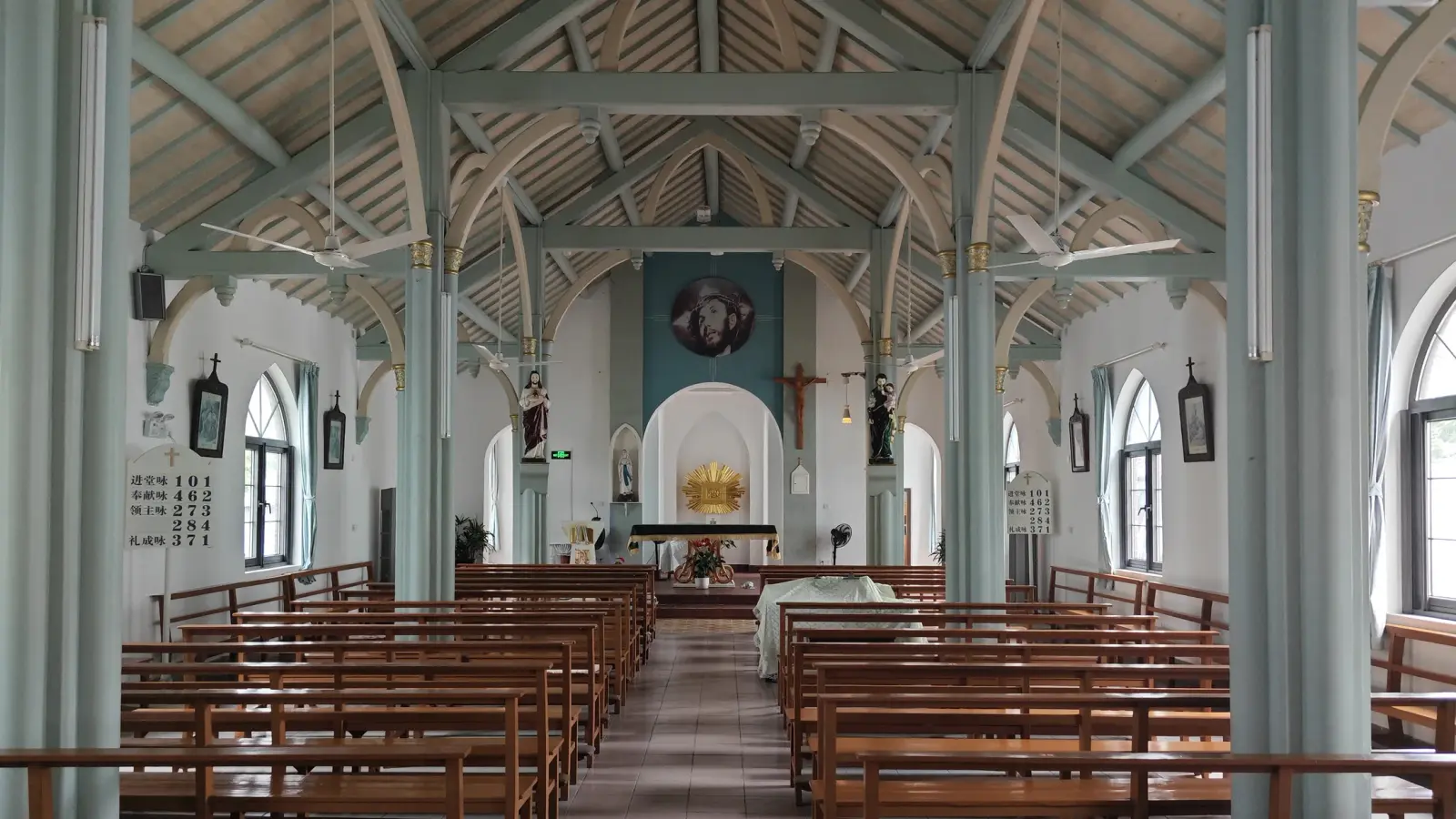 A church interior with mint-green iron columns, exposed roof trusses, wooden benches and a small apse with a crucifix.