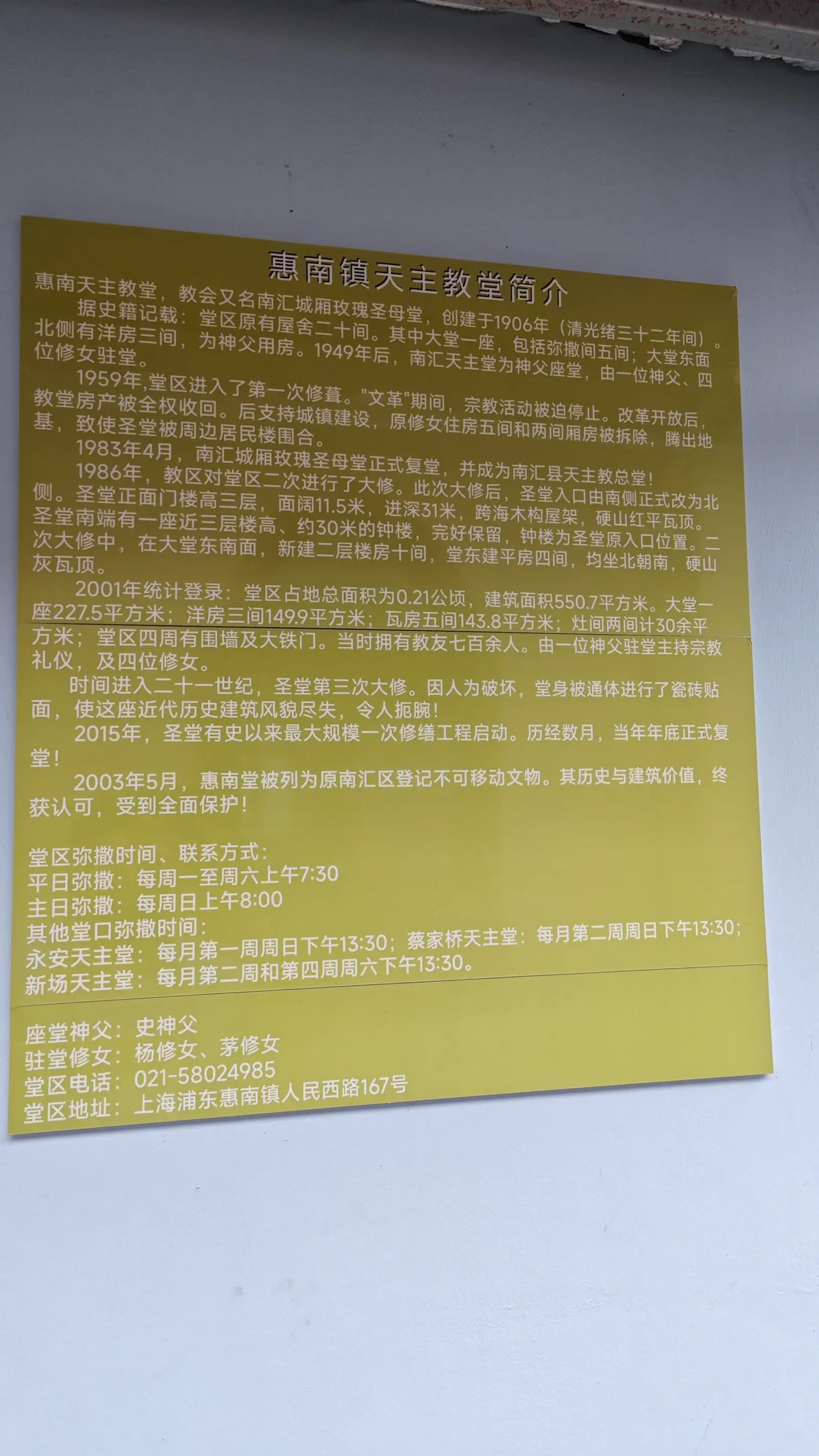 A yellow board with dense Chinese text on a white wall, a history of the church and its Mass times.