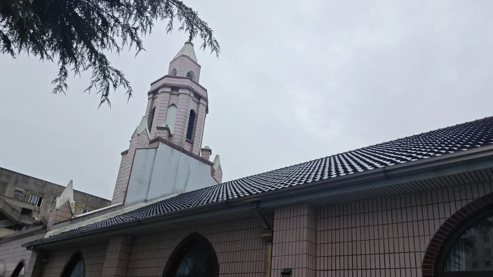 A pink-brick bell tower and a black tiled roof above a wall of pointed-arch windows, seen from below.