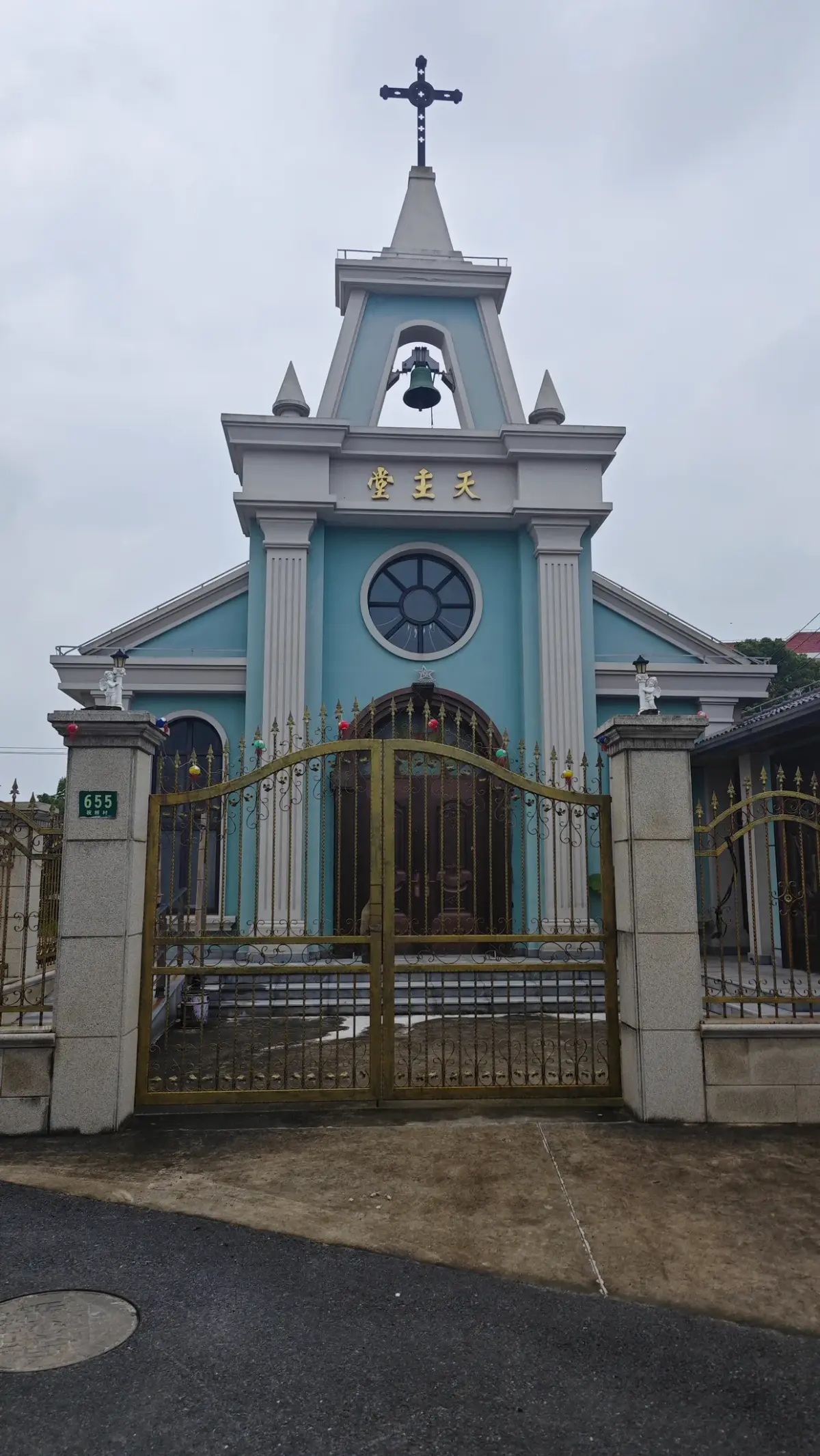 A pale blue church facade with a gabled bell tower, a round window and a cross, behind an ornamental metal gate.
