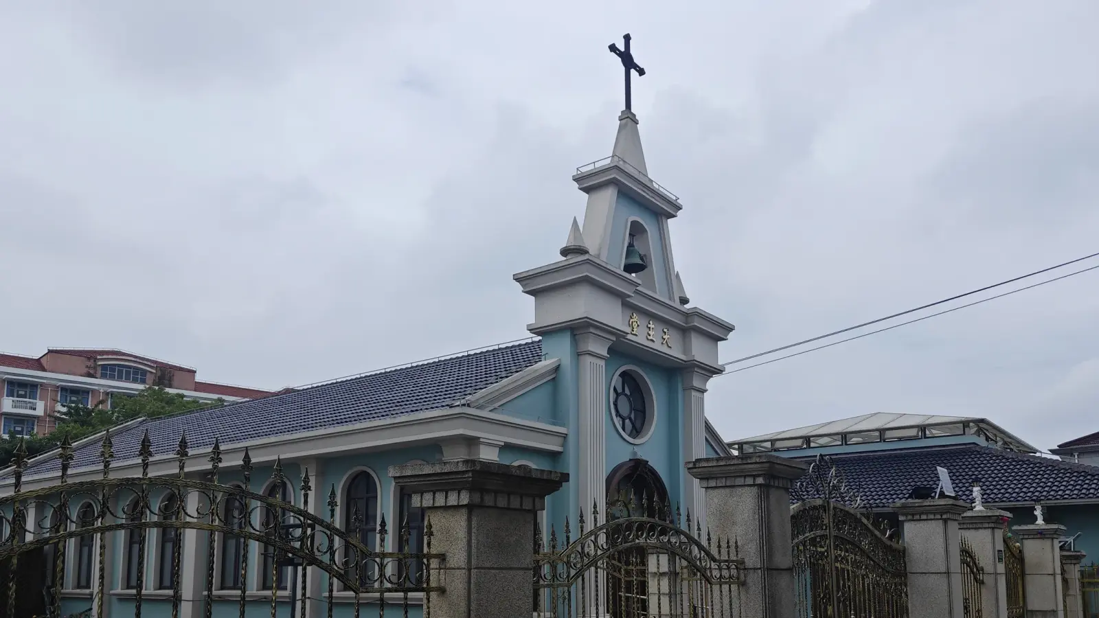 A blue church with a tall gabled bell tower and cross above a decorative iron fence and a grey tiled roof.