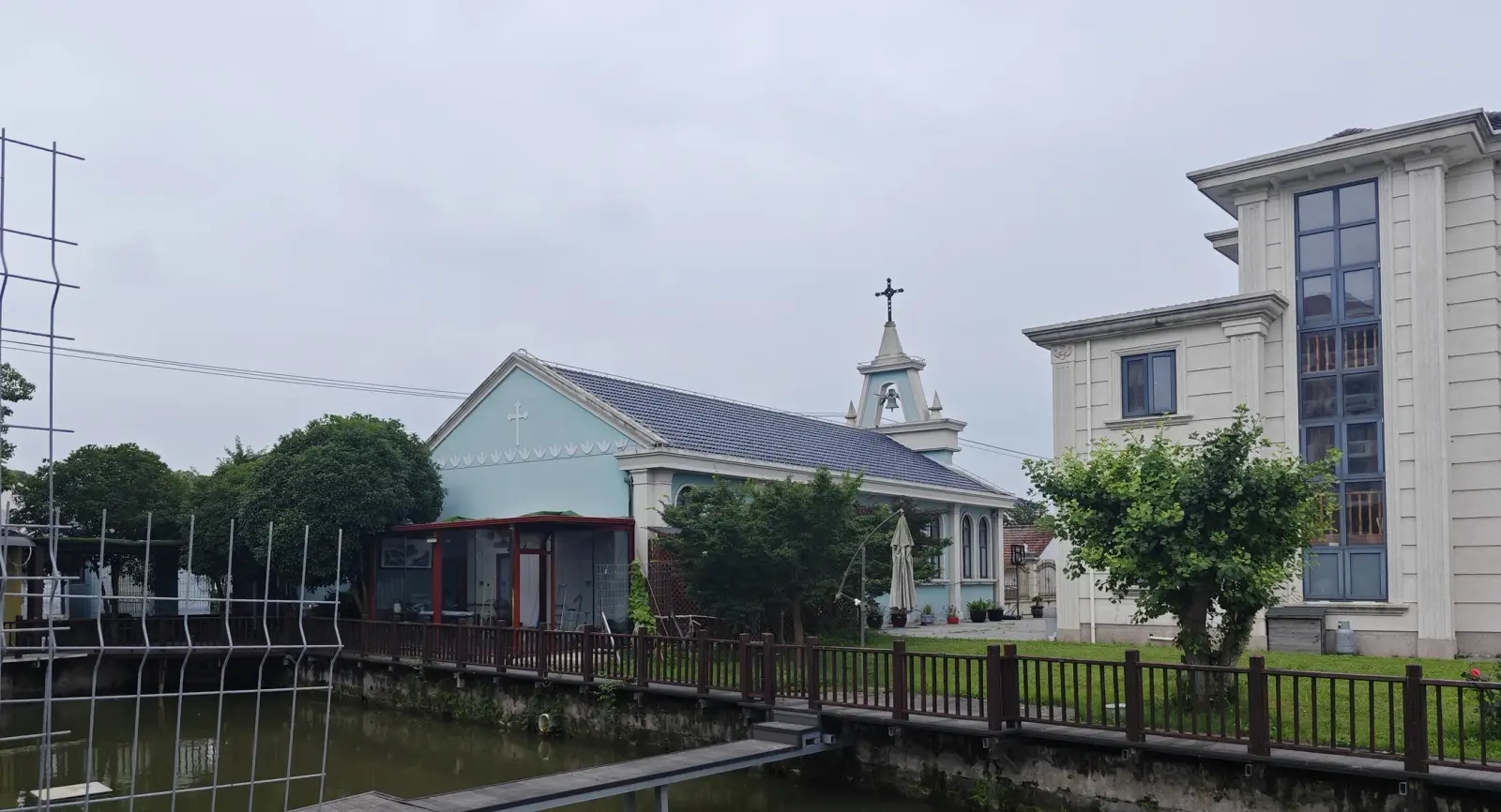 A small blue church with a small tower beside a pond and a large cream house under an overcast sky.