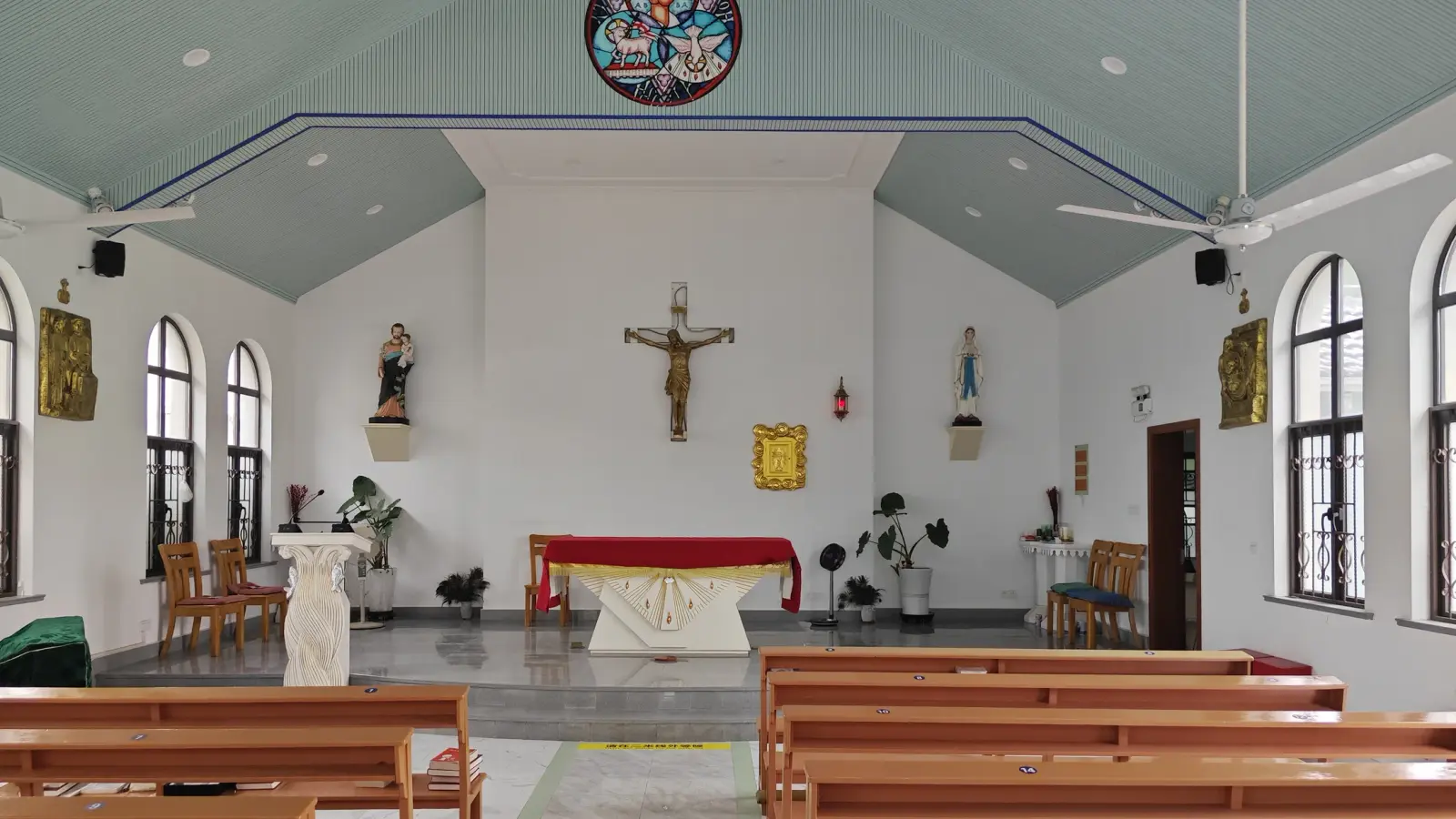 A bright church interior with a crucifix above a red-draped altar, two statues, arched windows and wooden benches.