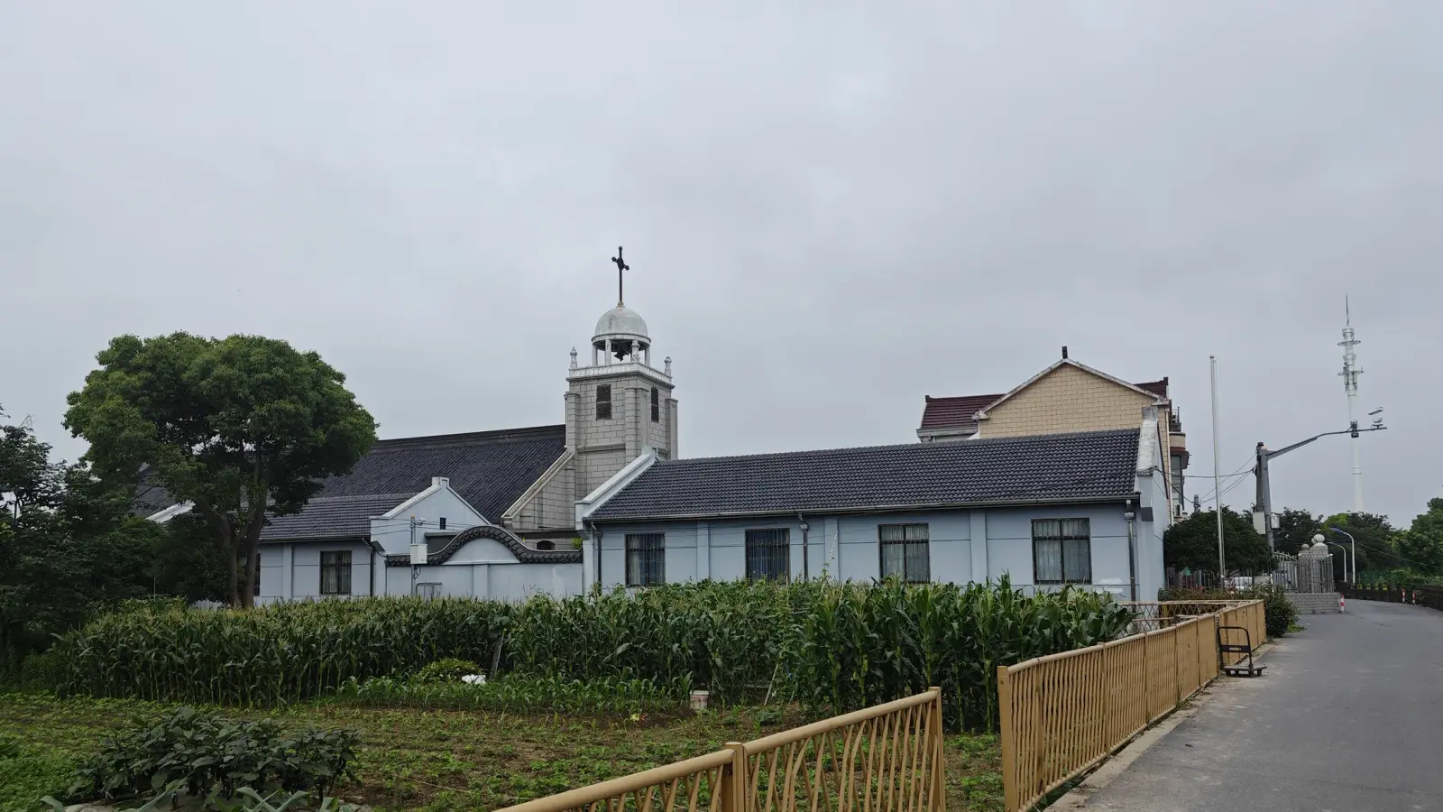 A long low blue-grey church building with a small tower and cross, beyond a field of crops and a yellow railing.