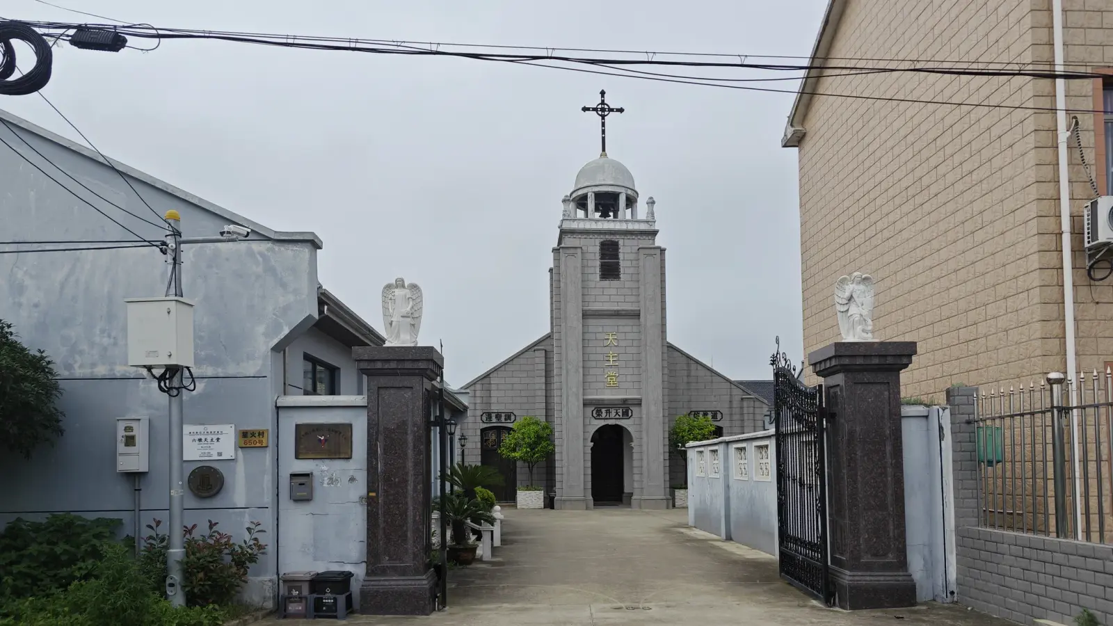 A narrow courtyard framed by gate posts leads to a grey church front with a central tower topped by a cross.