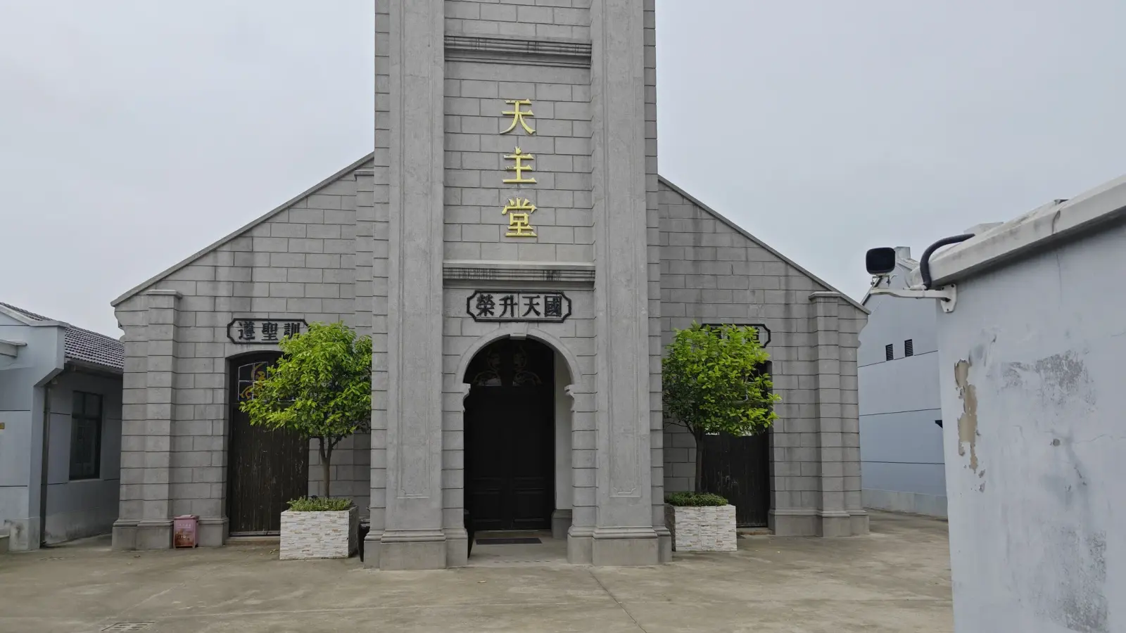 Close view of a grey church facade with a tall central tower, an arched black door and gold characters on the tower.