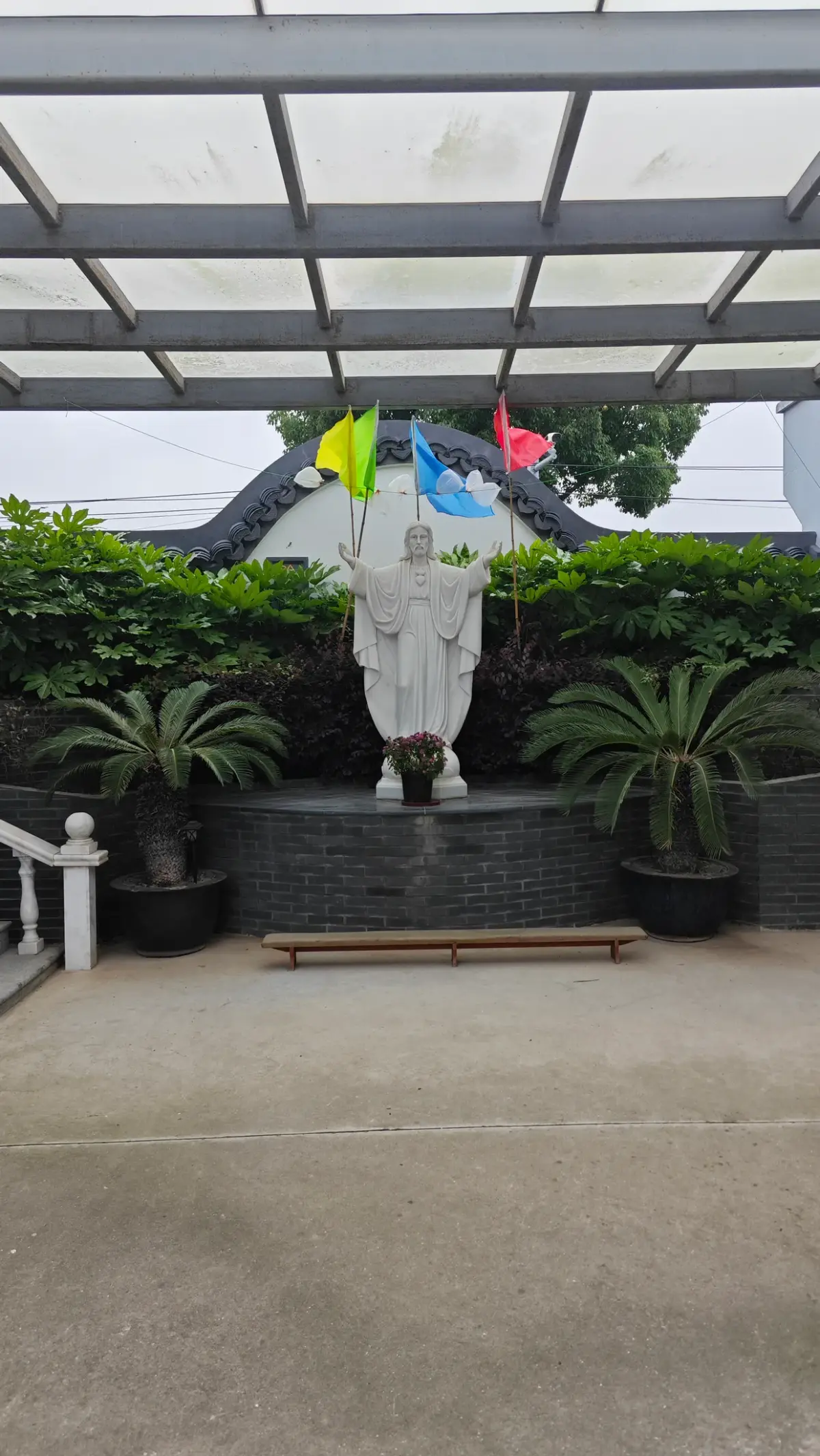 A white statue of a robed figure with arms outstretched, set among potted plants under a translucent canopy.