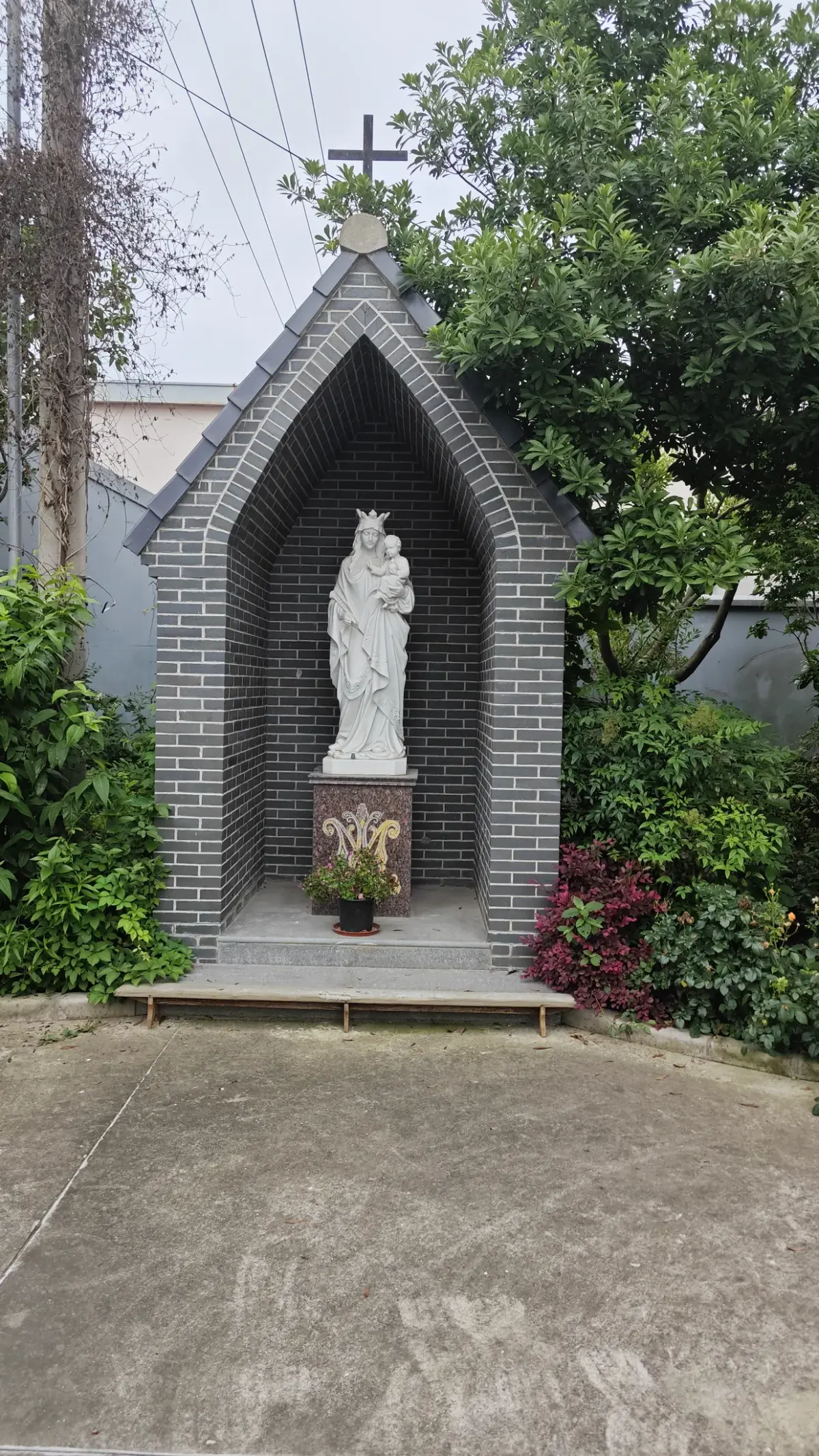 A white statue of a woman holding a child inside a pointed grey brick niche surrounded by greenery.