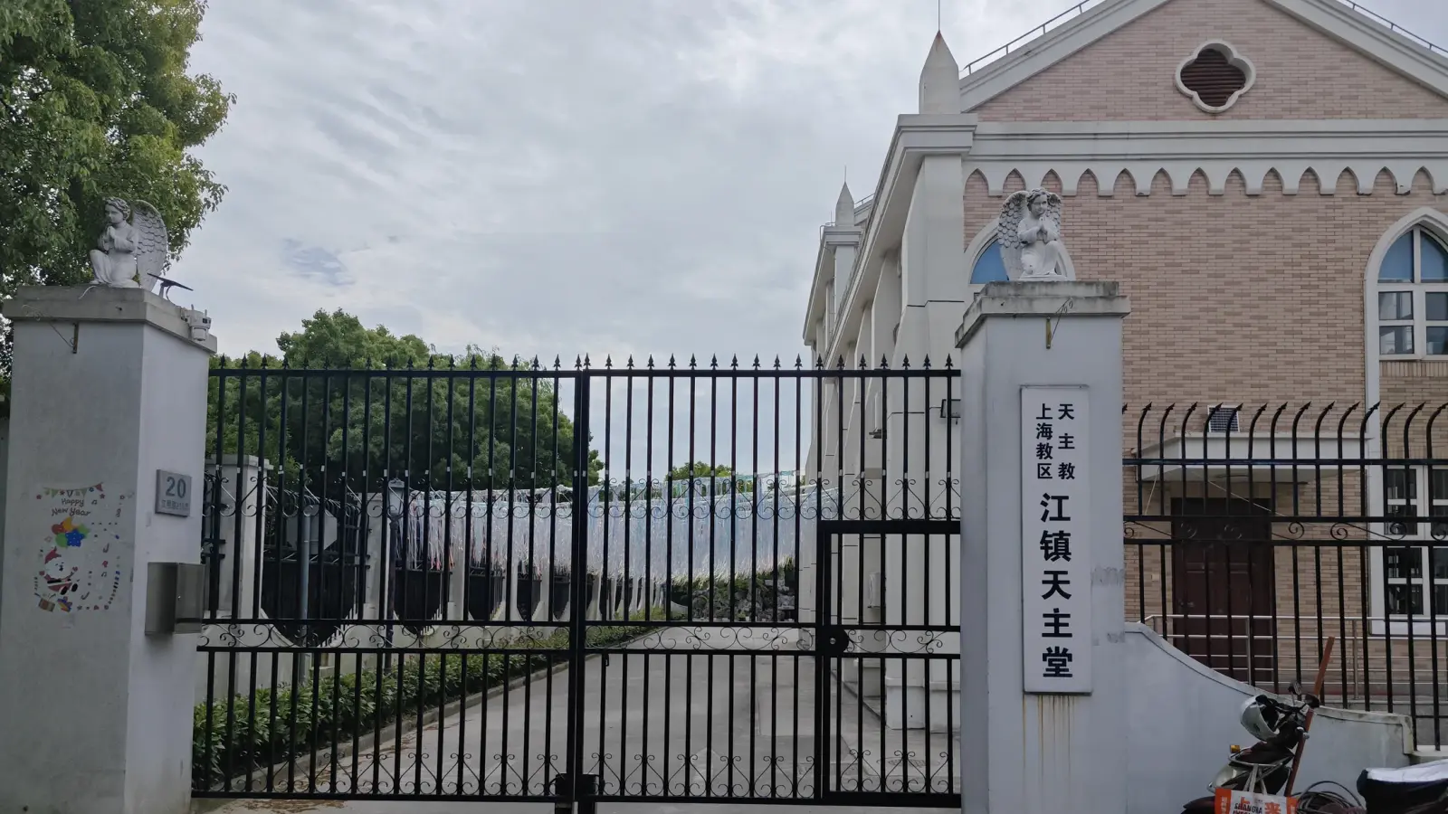 A black iron gate between white gate posts, with a vertical white name board in Chinese, beside the pink-brick church.
