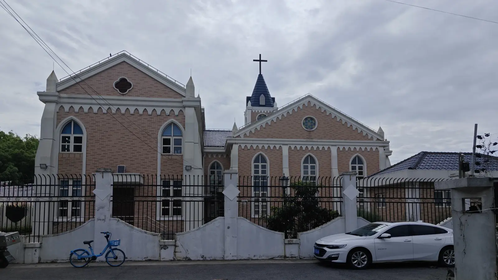 A pink brick church with white trim, pointed windows and a small blue-topped tower with a cross, behind a black iron fence.