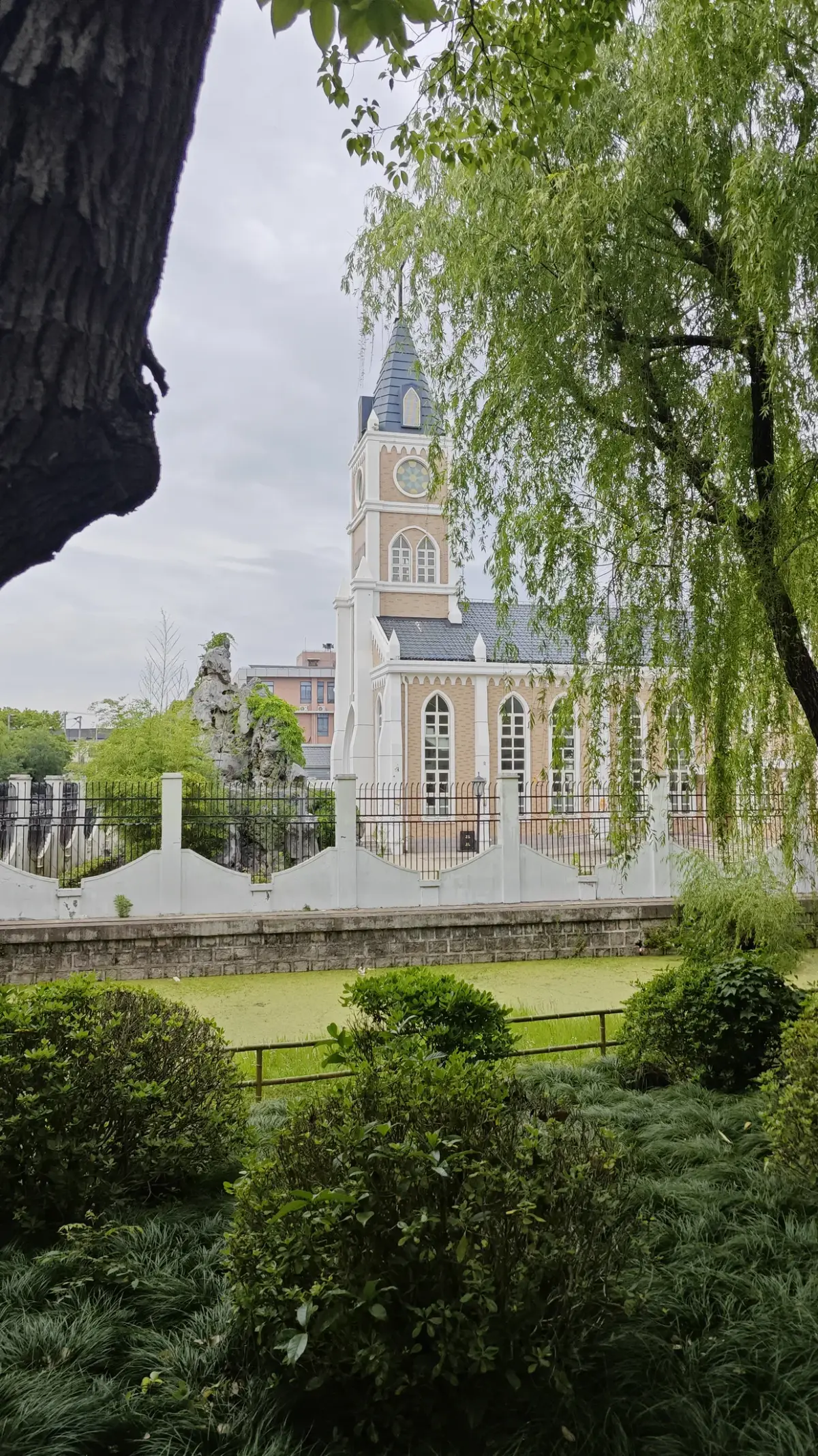 A church tower with a blue spire among trees, framed by willow branches, beyond a pond and hedge.