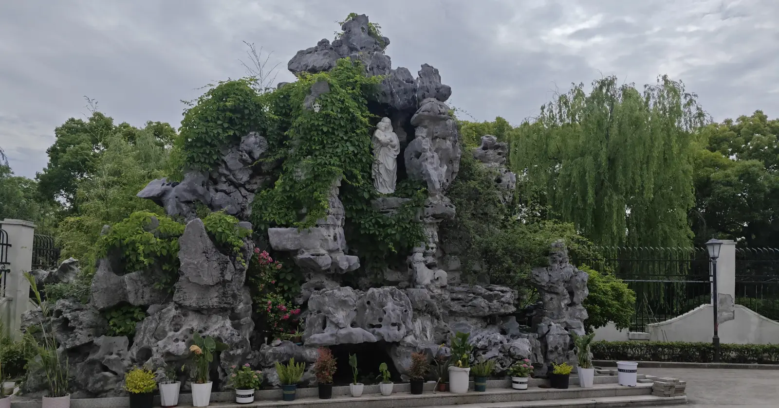 A mound of grey garden rocks covered with greenery and a white statue in a niche, with potted plants in front.