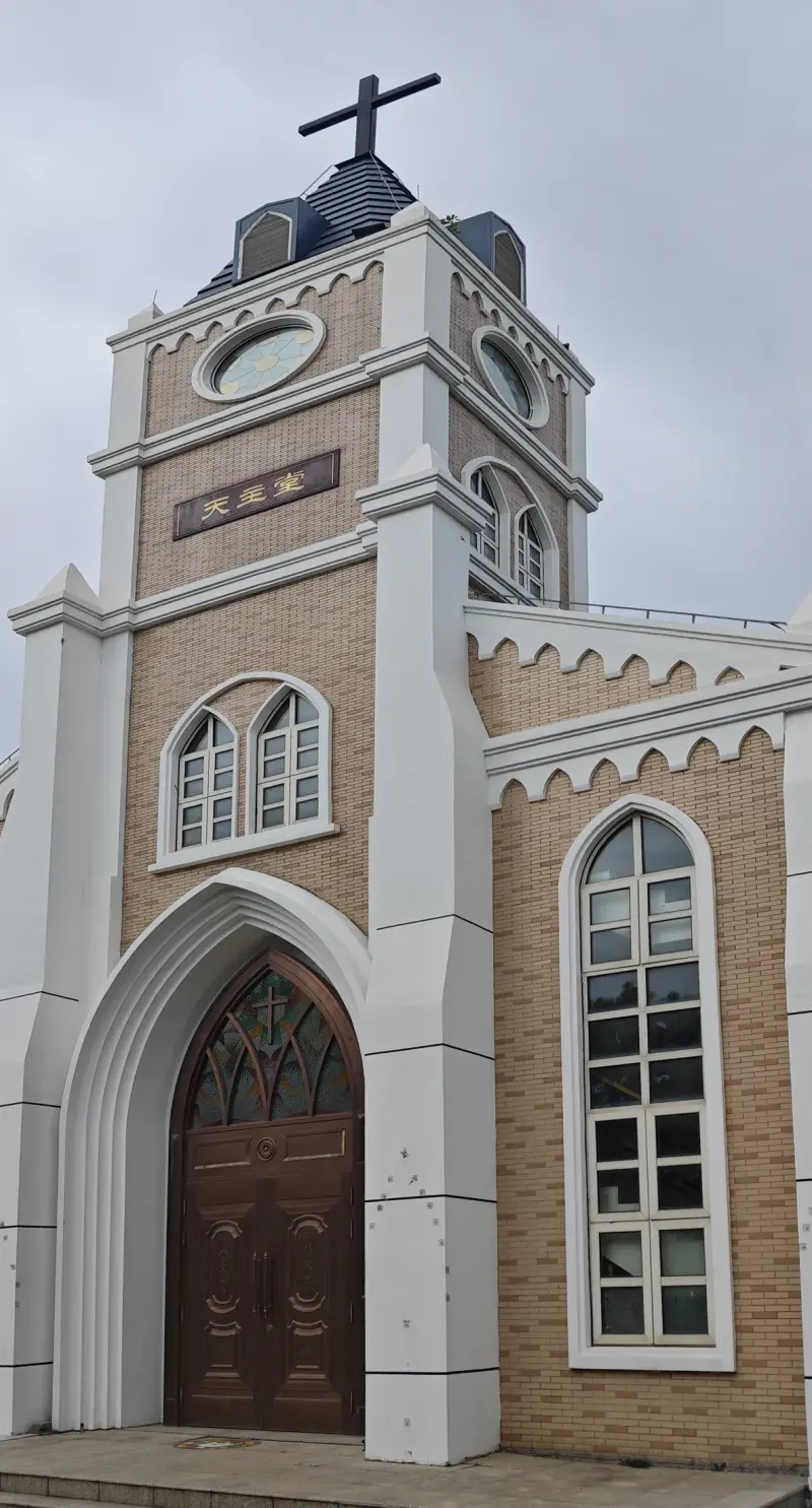 A pale brick tower with white buttresses, a clock, a cross, and a tall arched wooden door.