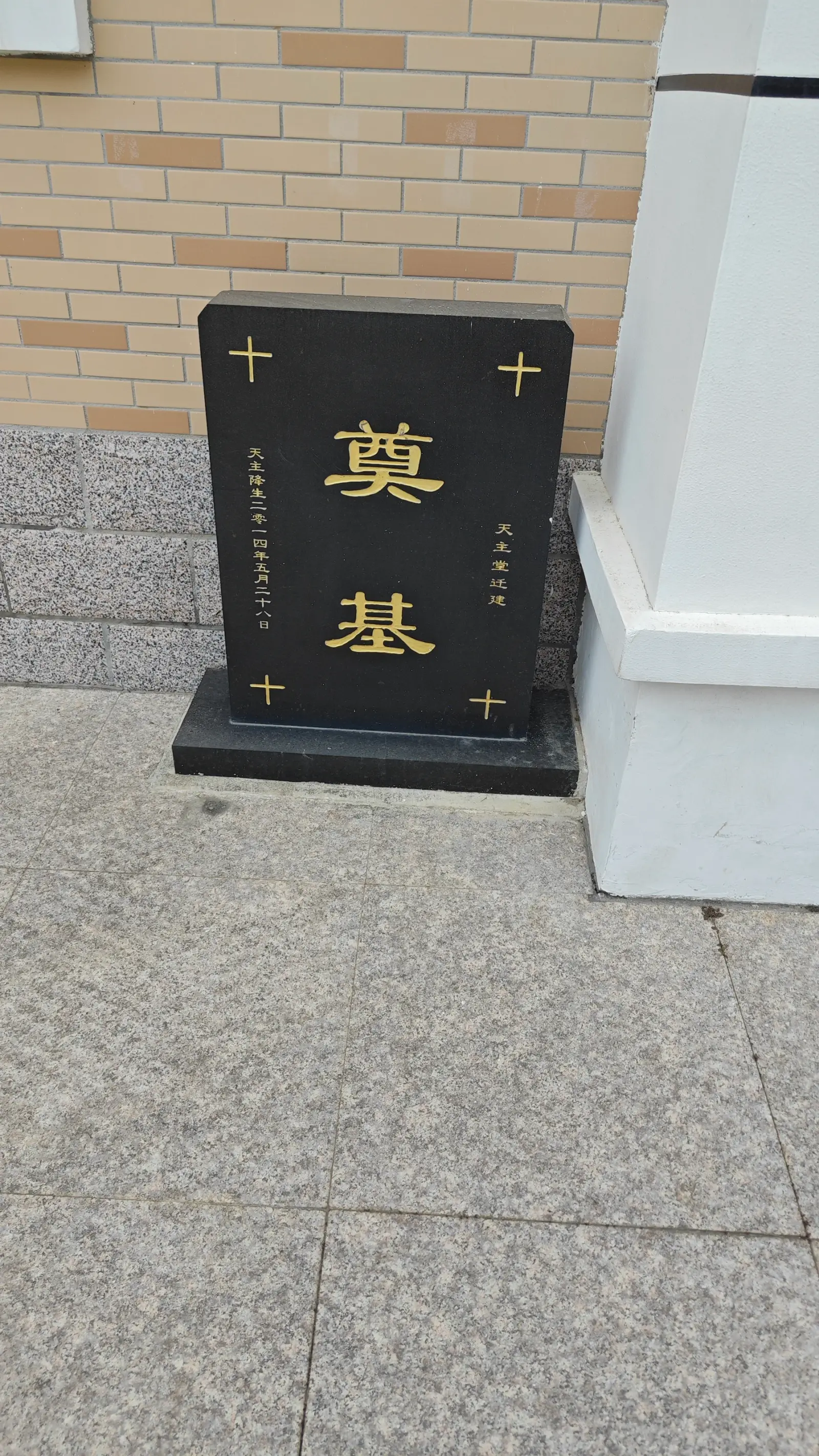 A black stone tablet with two gold Chinese characters and small crosses beside a white corner buttress.