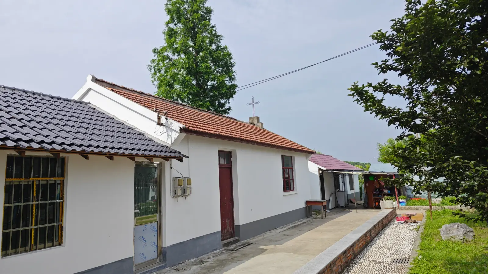 A single-storey white building with a red tile roof and a cross on the gable end beside a paved path and trees.