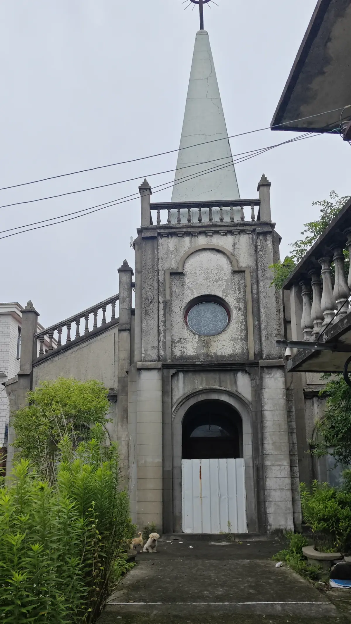 A narrow stone tower with a pointed spire and cross above a round window and an arched white door, with a path leading to it.