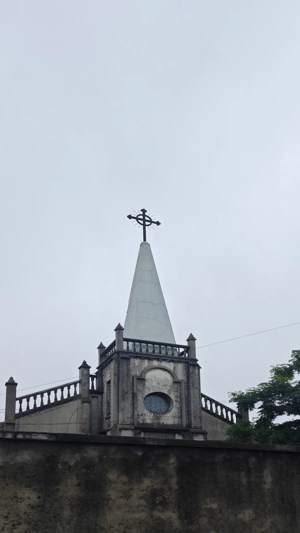 A grey spire with a cross above a small balustrade and round window, behind a dark wall.