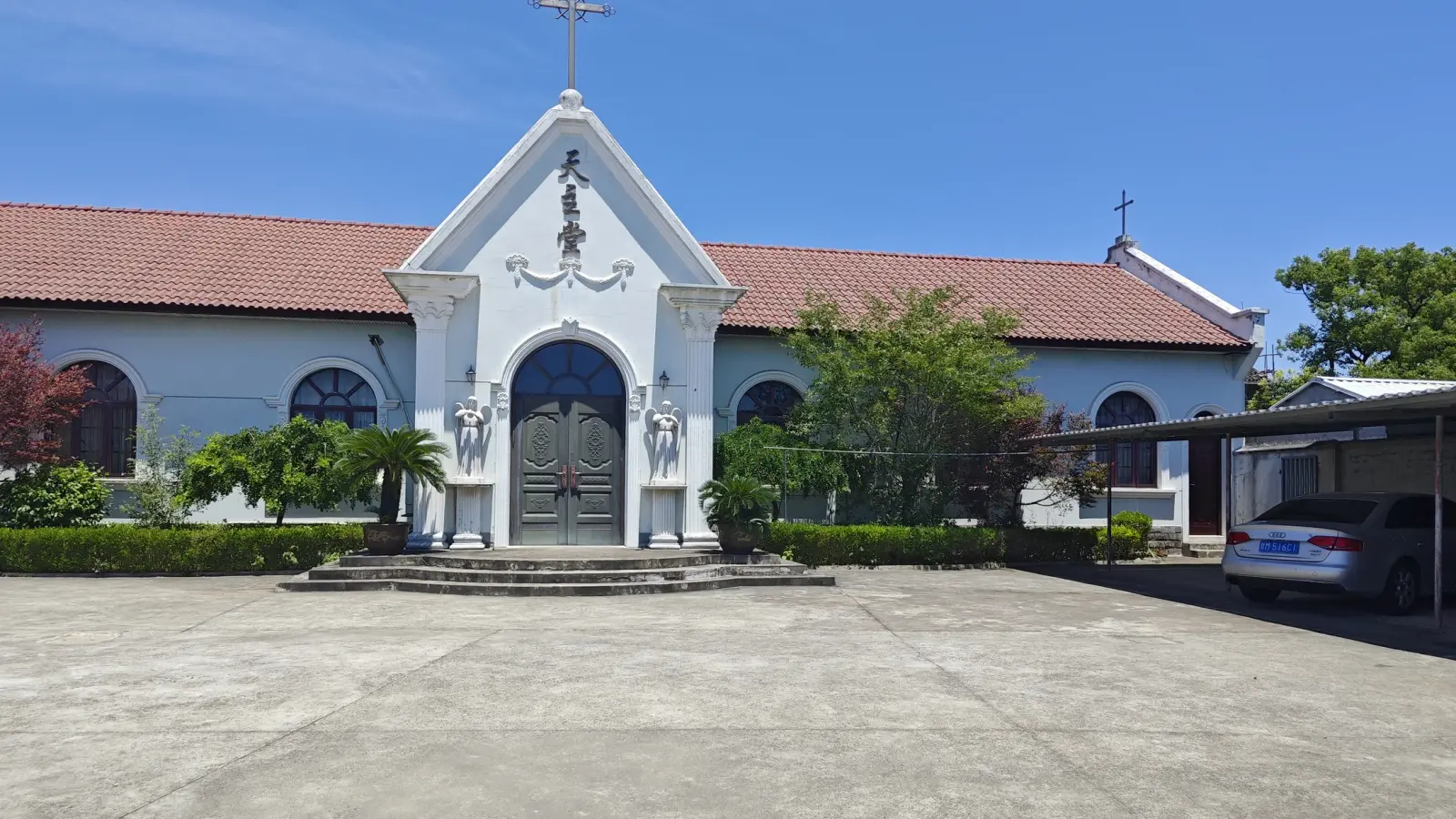 A white church front with a pointed pediment, a cross and a carved panel above an arched grey door, with red tiled wings either side.