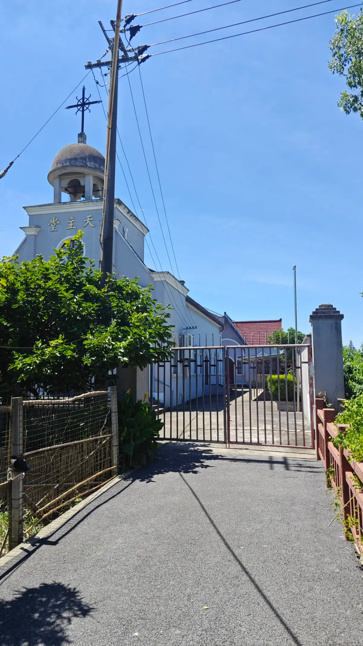 A narrow tarmac lane leading to an iron gate, with a white church and domed belfry behind and a utility pole at left.