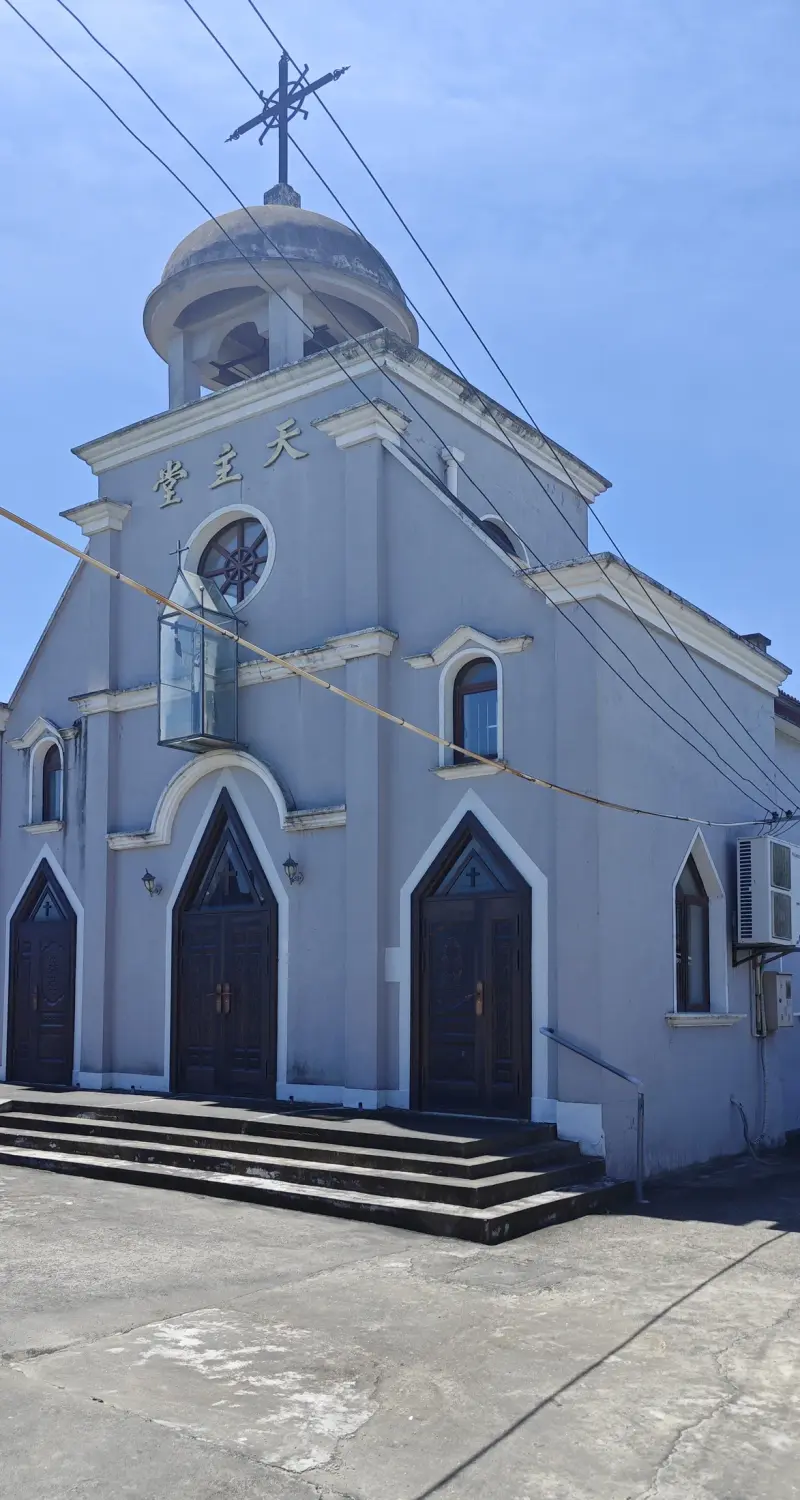 A pale grey-blue church front with three pointed arched wooden doors, a round window and a small domed belfry with a cross, under a clear sky.