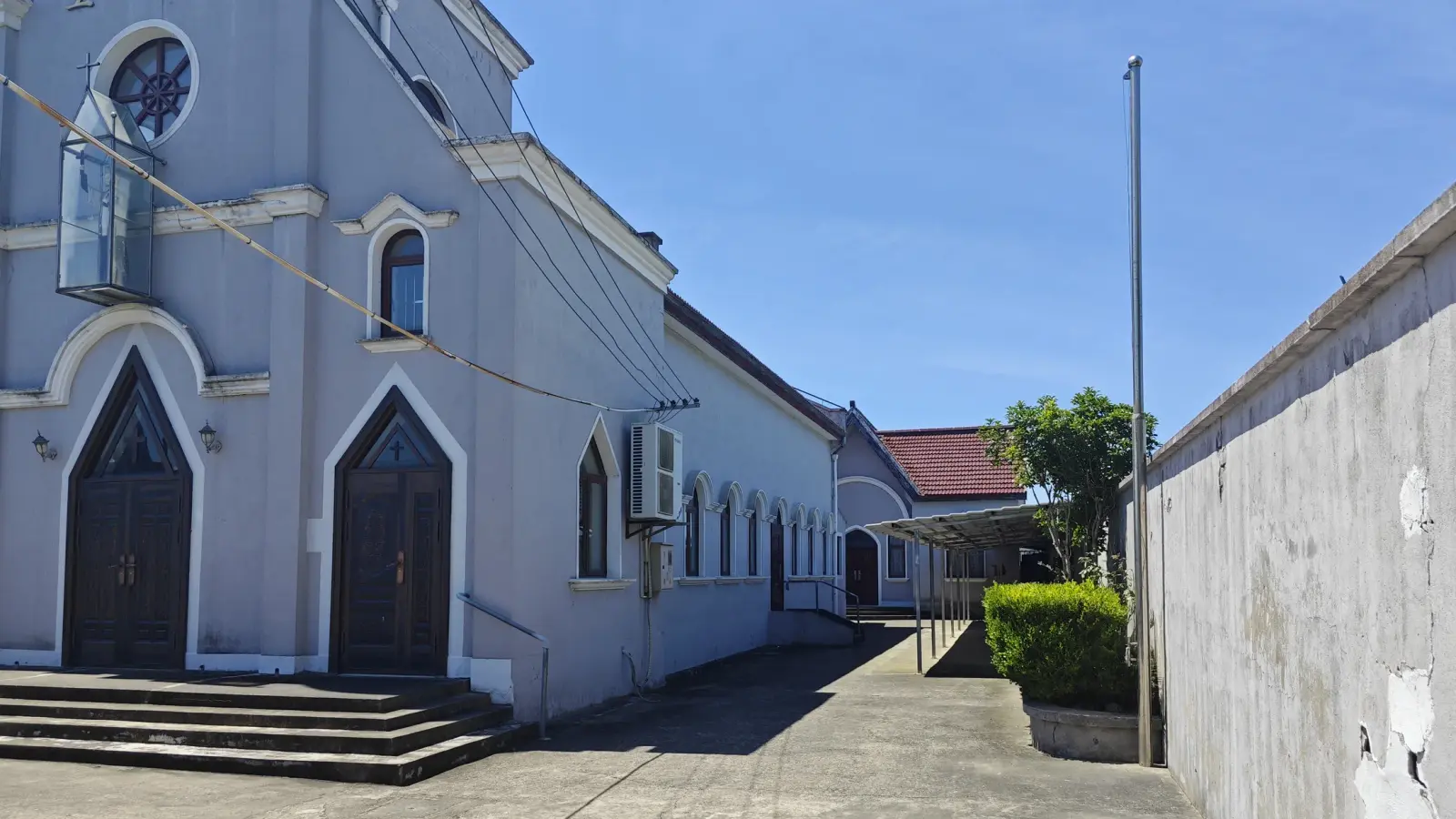 A pale church front and side wall with pointed arched doors, stone steps and a row of small arched windows beside a concrete wall.