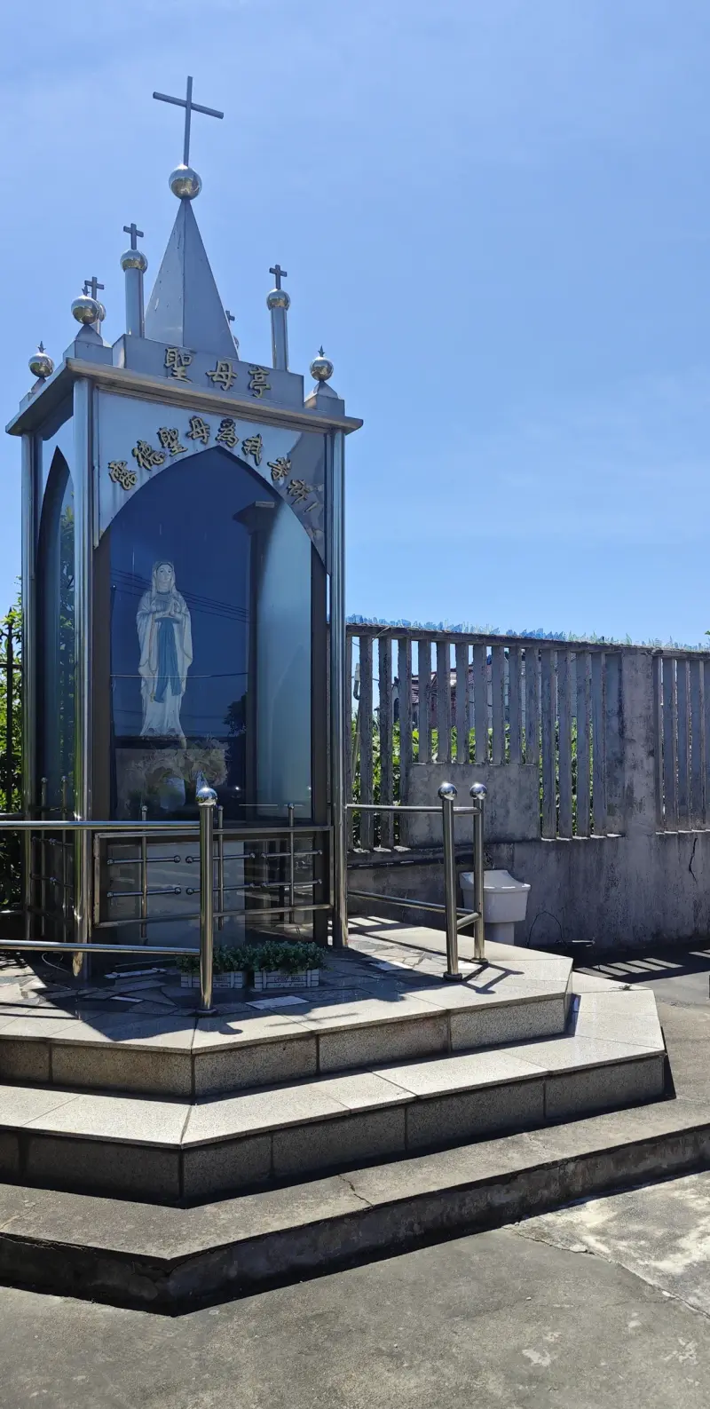 A hexagonal blue and white shrine with a pointed top and cross, sheltering a white statue, with railings around its stepped base.