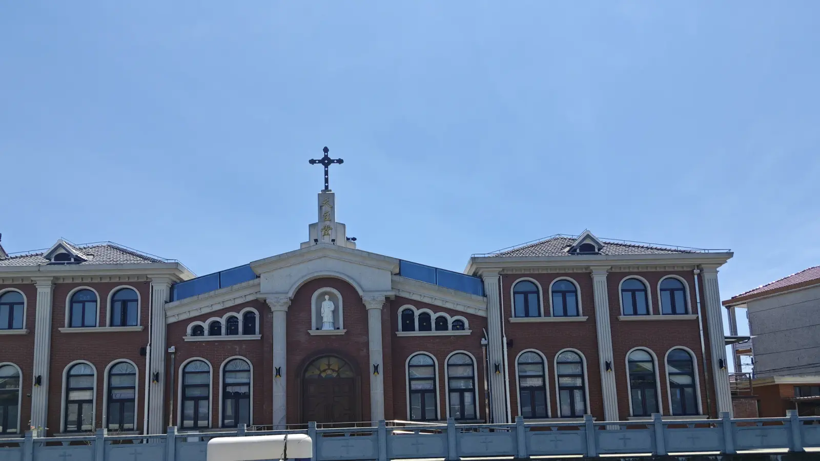Looking up at a brick church front with a central pediment, a cross, a statue in a niche and rows of arched windows.