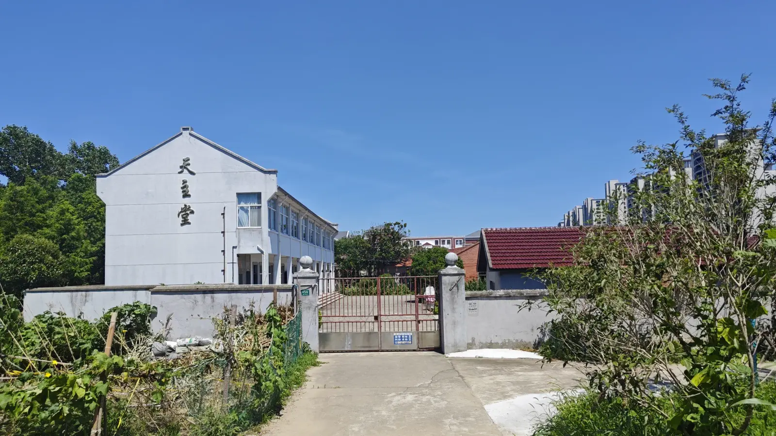 A white building with Chinese characters on its gable end behind a wall and a closed iron gate, with trees and apartment blocks beyond.