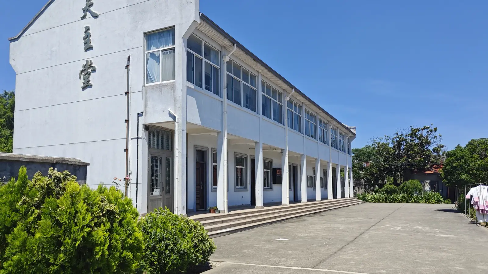 A white two-storey building with a tall gable bearing large Chinese characters and a long colonnaded verandah beside a paved yard.