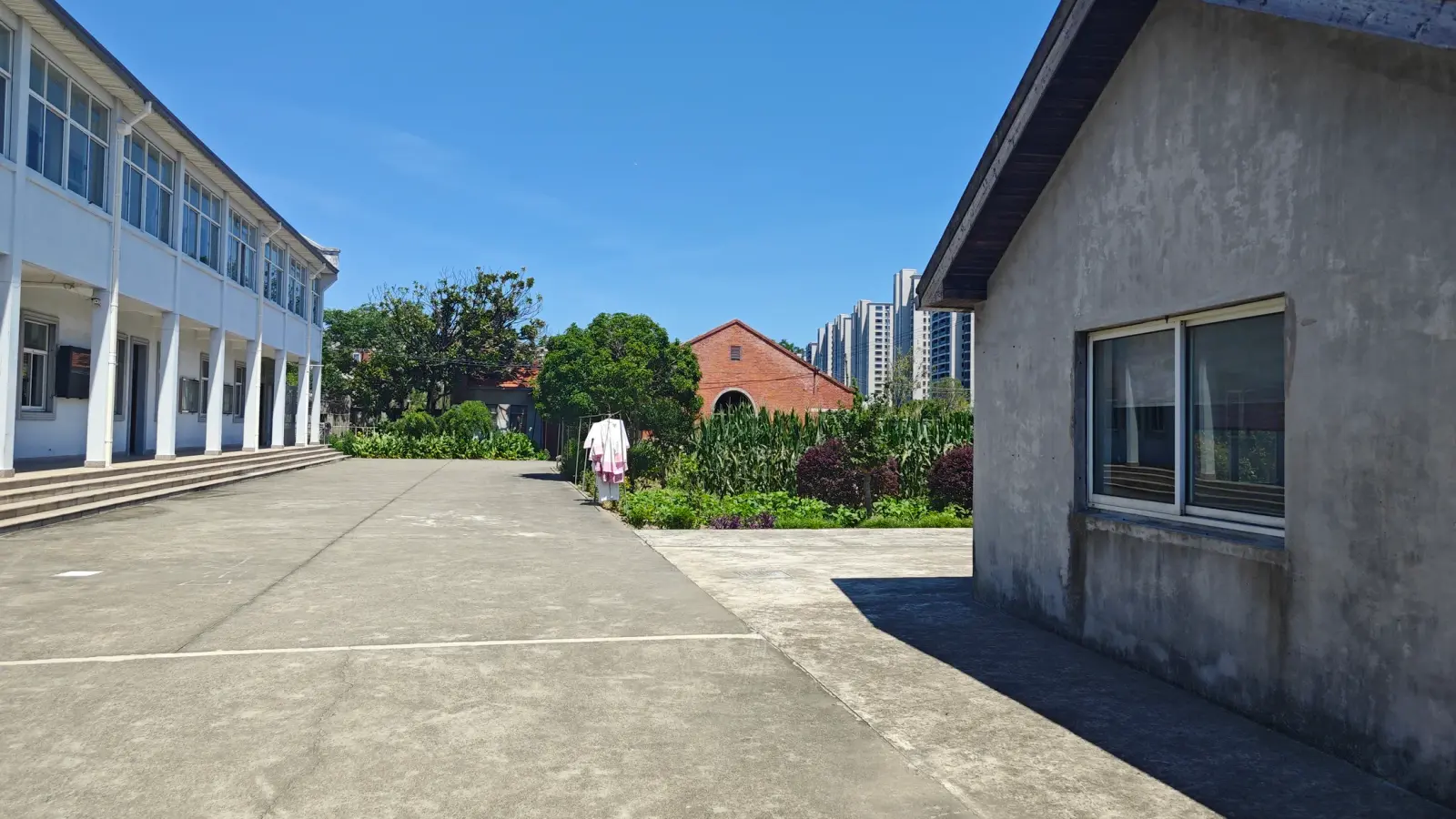 A wide paved courtyard beside a white two-storey building with a long row of windows, and a grey outbuilding to the right.